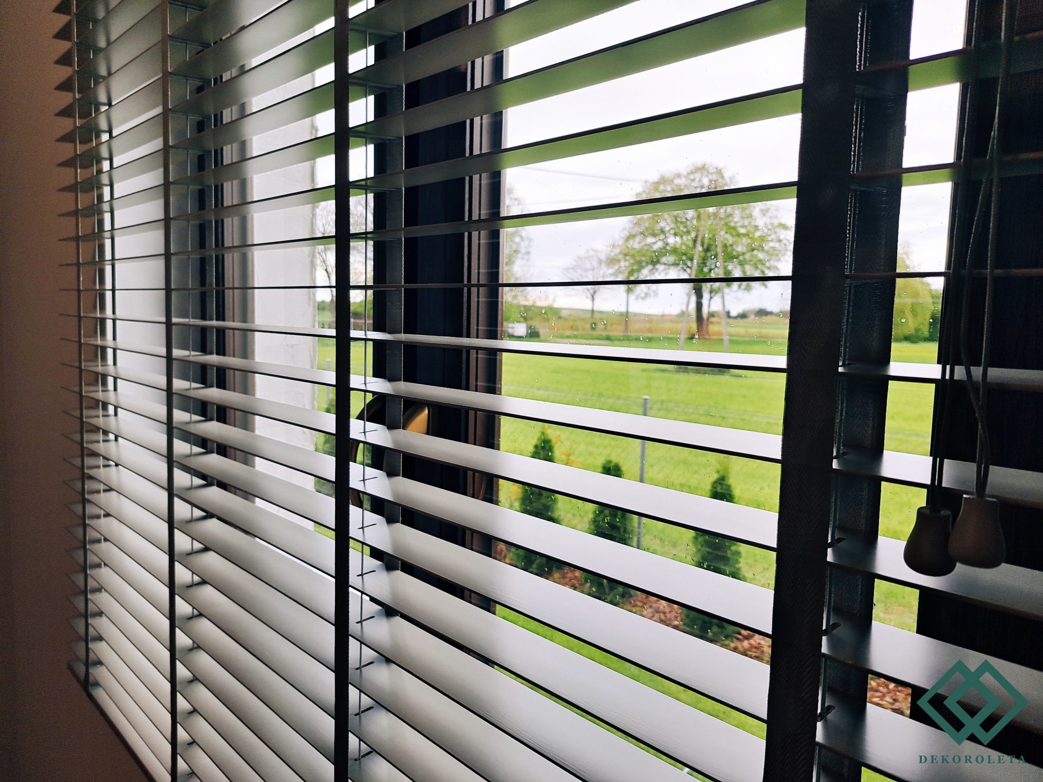 Zamknięte białe żaluzje drewniane z widokiem na zielony ogród w deszczowy dzień, z kroplami wody na szybie.