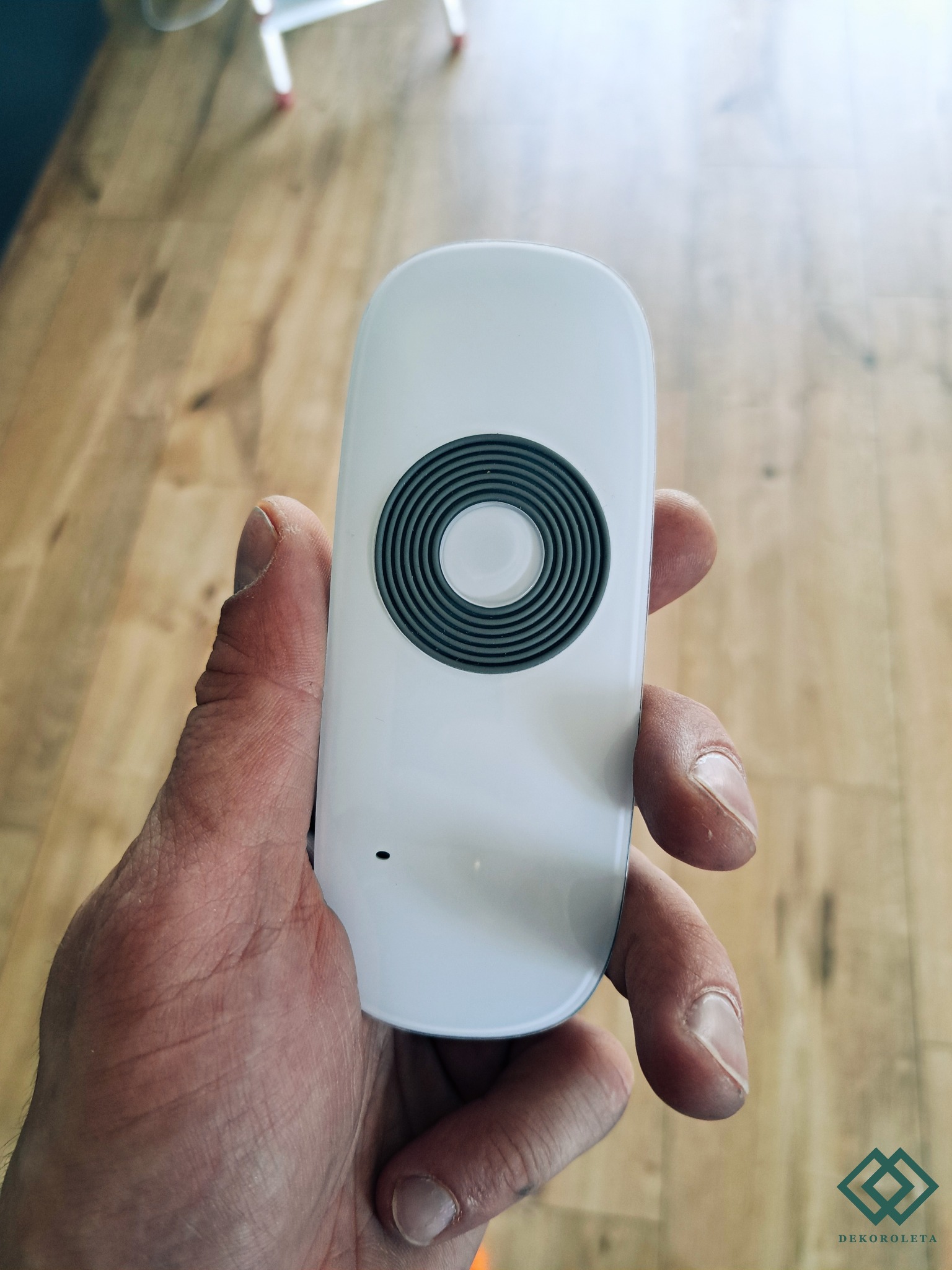 Biały pilot do sterowania roletami z szarym okrągłym przyciskiem w dłoni na tle podłogi z paneli.