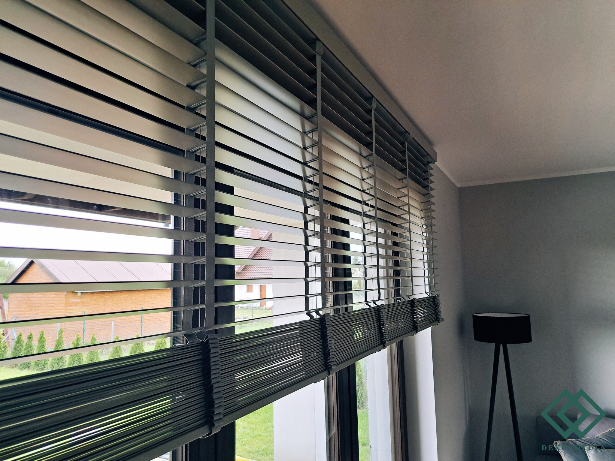 Szare poziome lamele w oknie, widok na ogród i drewniany dom, minimalistyczna lampa w tle.