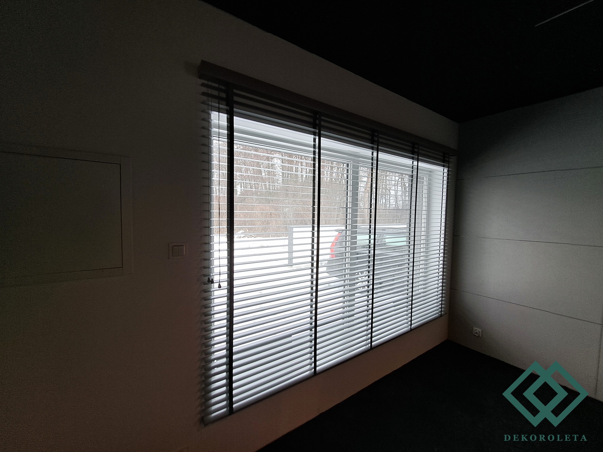 Zamknięte poziome białe listwy w oknie z widokiem na zimowy krajobraz, widoczny fragment logo firmy w prawym dolnym rogu