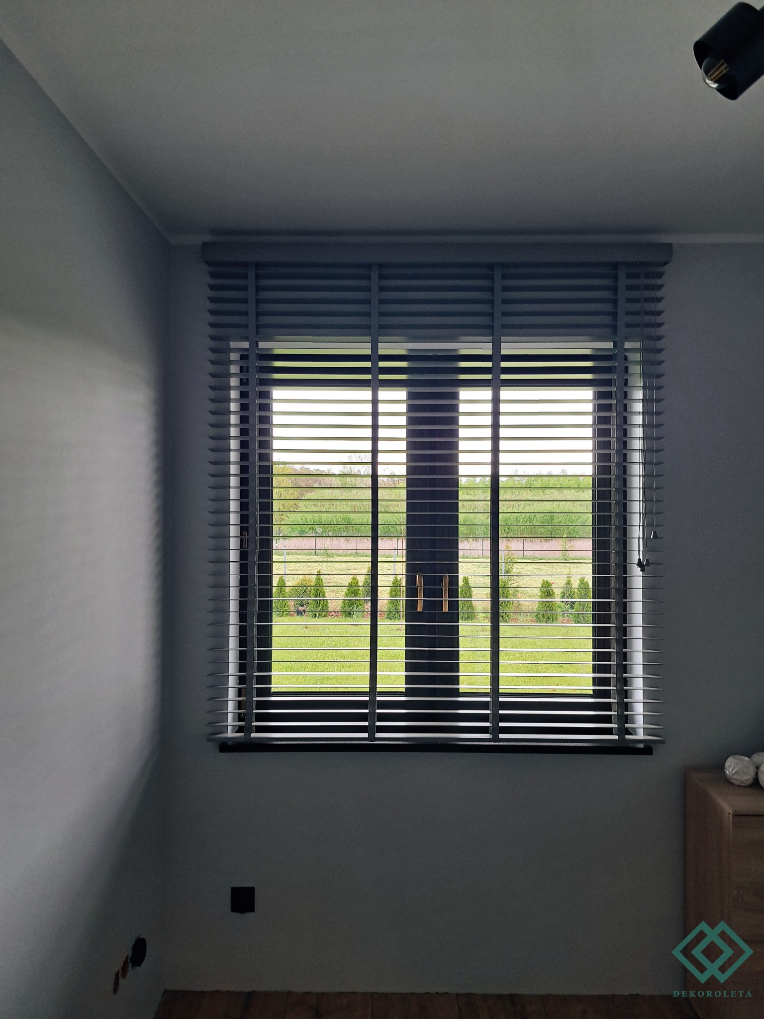 Szare poziome osłony okienne z drewnianymi lamelami, widok na zielony ogród z tujami, szare ściany i drewniana podłoga w pokoju.