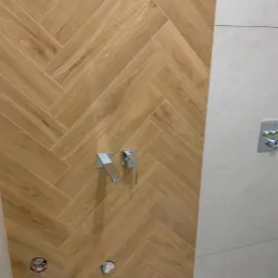 Fragment ściany łazienki z ułożonymi w jodełkę płytkami imitującymi drewno, chromowana bateria umywalkowa, otwory na podłączenie armatury.