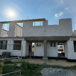 Surowy stan domu jednorodzinnego z białych bloczk&oacute;w silikatowych, widoczne pakiety materiał&oacute;w budowlanych na g&oacute;rze, ogrodzenie tymczasowe z siatki, słoneczny dzień.