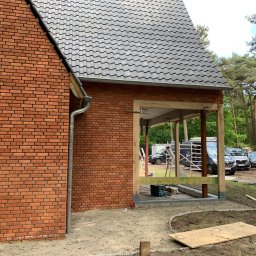RD Construction Radosław Dyc - Częściowy widok murowanego domu z cegły z dobudowywaną drewnianą konstrukcją zadaszenia; widoczne narzędzia i samochody w tle.