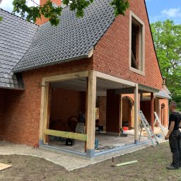 RD Construction Radosław Dyc - Budowa domu z czerwonej cegły z widocznym fragmentem dachu z szarej dachówki, drewnianą konstrukcją werandy i pracownikiem obserwującym postęp prac.