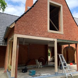 RD Construction Radosław Dyc - Budowa domu z czerwonej cegły w trakcie realizacji, widoczne drewniane podpory i rusztowanie, fragment dachu z szarej dachówki, drabina oparta o ścianę, narzędzia budowlane w tle, nieukończony komin.