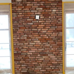 RD Construction Radosław Dyc - Ściana z czerwonej cegły dekoracyjnej z otworem na instalację, zabezpieczona taśmą malarską przy oknach. Widoczny fragment podłogi i rura kanalizacyjna.