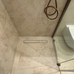 RD Construction Radosław Dyc - Beżowa kabina prysznicowa z odpływem liniowym i miedzianą armaturą, widok z góry. Uwagę zwraca dopasowanie kolorystyczne płytek i elementów wykończeniowych.