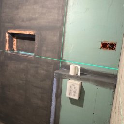 RD Construction Radosław Dyc - Niedokończona łazienka z surowymi ścianami, zielonymi płytami gipsowymi, laserem krzyżowym, wnęką i częściowo widoczną muszlą klozetową. Etap prac wykończeniowych.