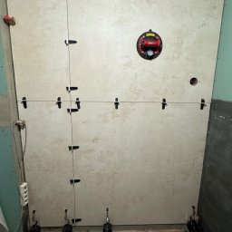 RD Construction Radosław Dyc - Układanie beżowych płytek na ścianie z użyciem systemu poziomowania. Widoczne przyssawki, krzyżyki dystansowe i poziomica laserowa. Etap prac wykończeniowych w łazience.