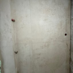 RD Construction Radosław Dyc - Wykończona ściana w łazience z beżowymi płytkami imitującymi kamień, widoczne punkty instalacji wodnej i elektrycznej. Minimalistyczny design.