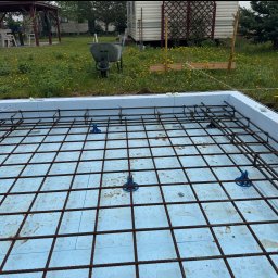 RD Construction Radosław Dyc - Zbrojenie fundamentu na styropianie z widocznymi podkładkami dystansowymi, przygotowane do zalania betonem. W tle taczka i teren budowy.