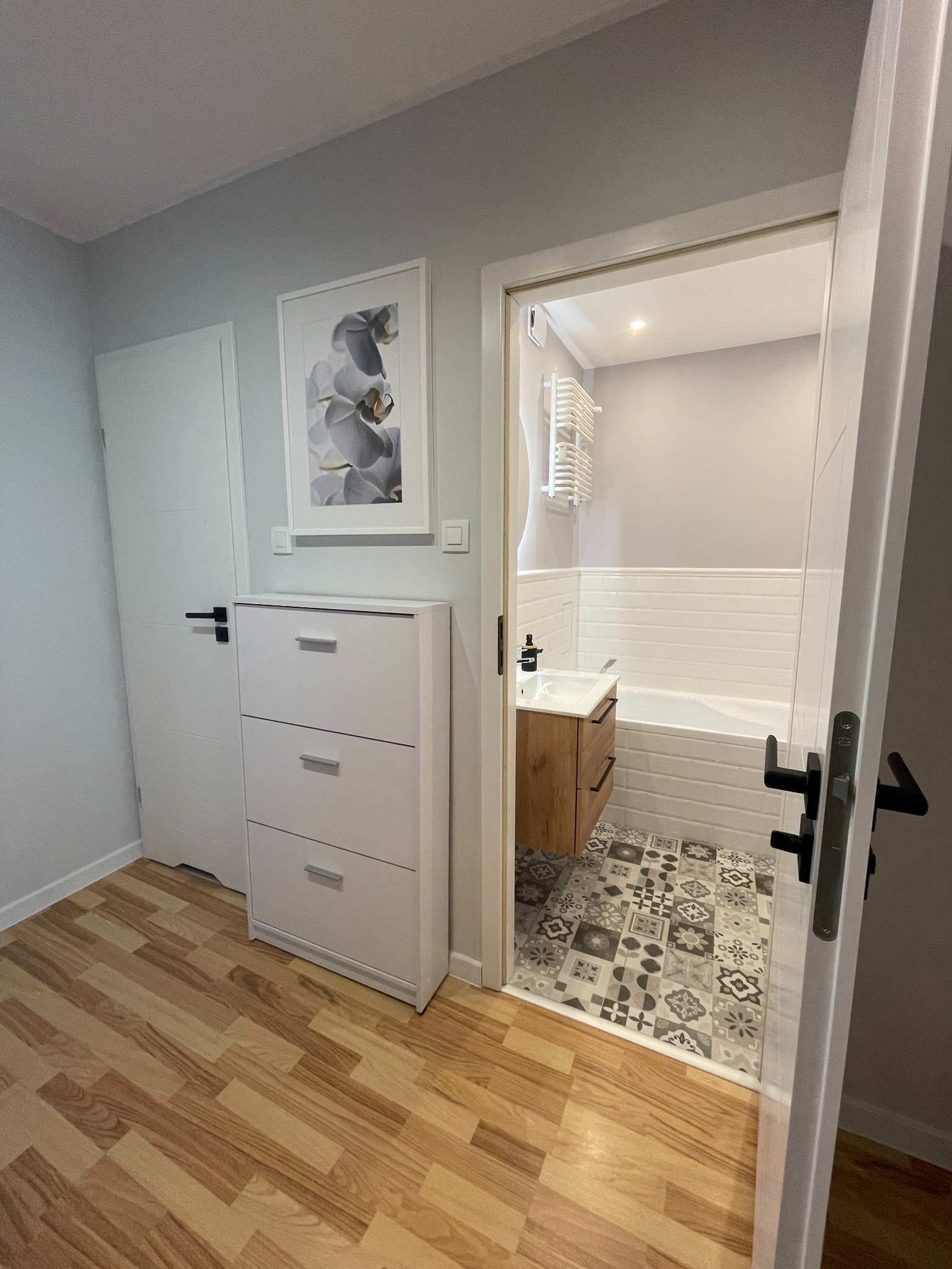 Widok na wykończoną łazienkę z wanną, umywalką na szafce z drewnianym frontem i grzejnikiem drabinkowym, przez otwarte drzwi z korytarza z drewnopodobną podłogą, białą szafką na buty i obrazem...