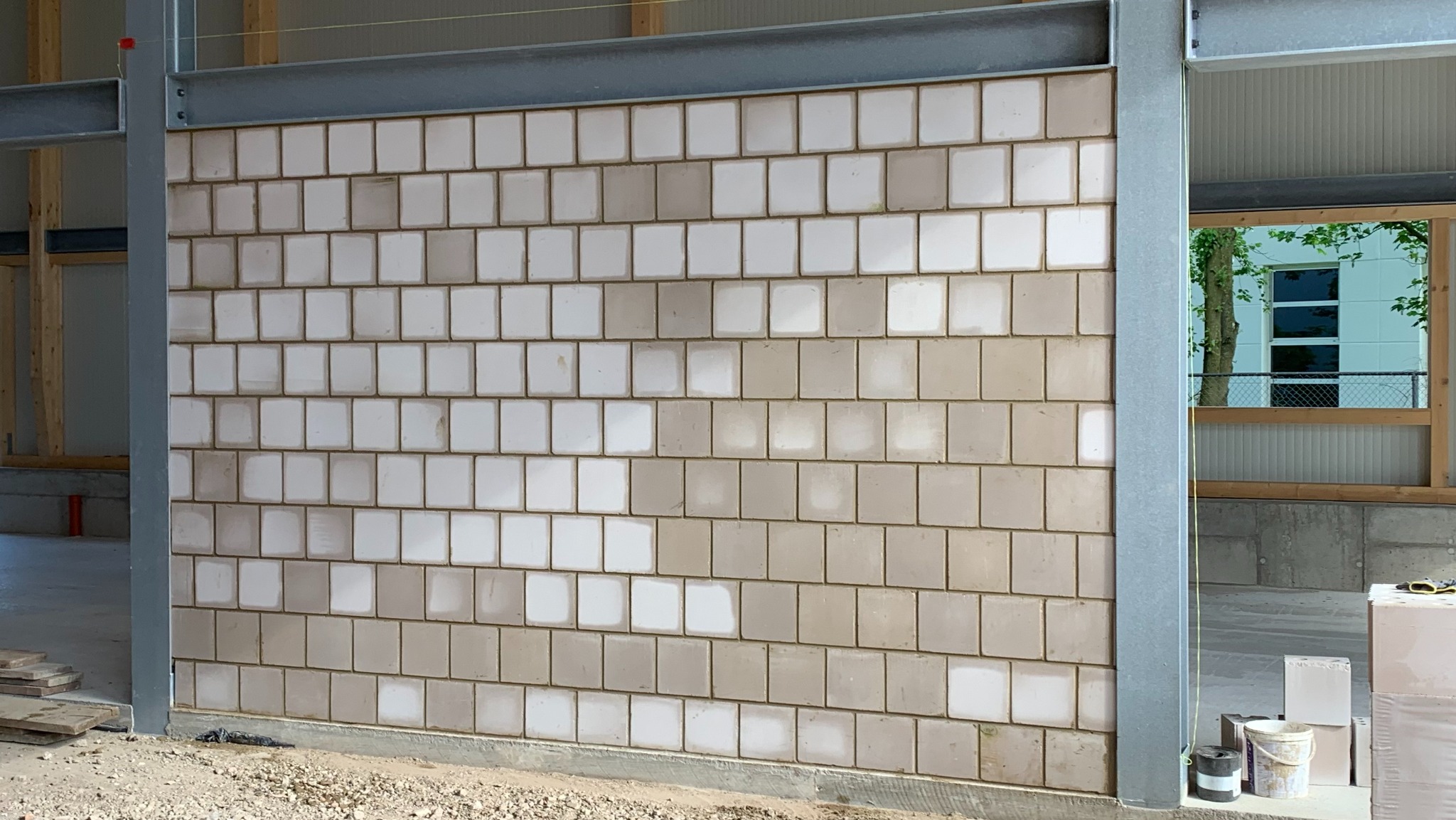 Ściana z białych, kwadratowych płytek ceramicznych, częściowo zabrudzonych, w surowym stanie, z widoczną stalową konstrukcją nośną budynku.