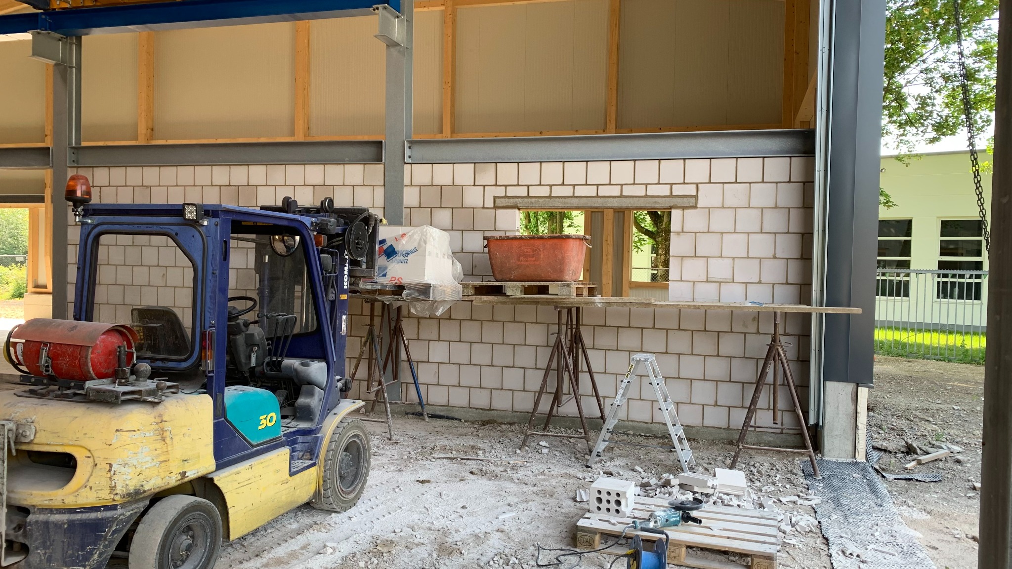 Wnętrze powstającego budynku murowanego z widocznym wózkiem widłowym, rusztowaniami podporowymi, pustakami i narzędziami budowlanymi, w tle budynek z oknami.