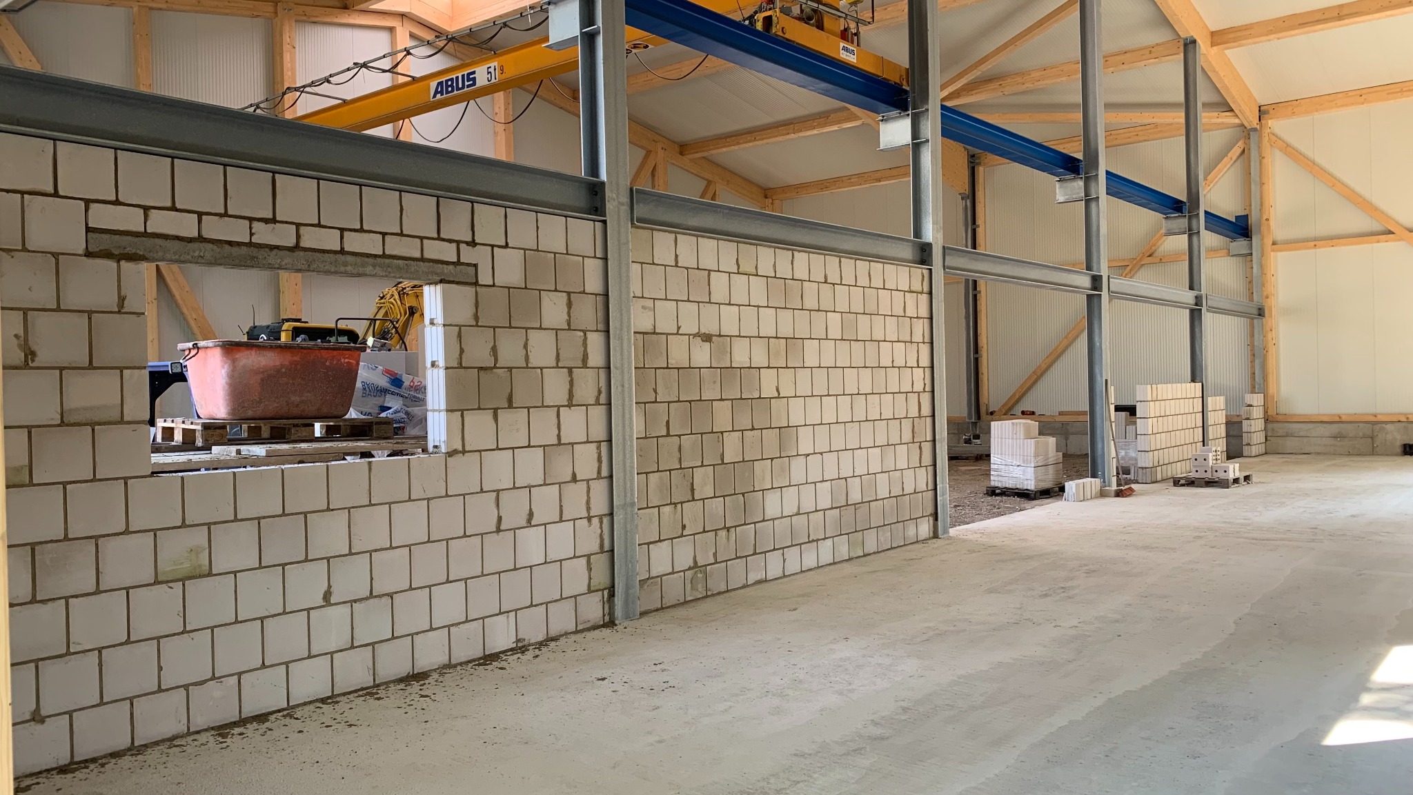 Wnętrze w trakcie budowy z bloczków, widoczne otwory okienne z paletami i pojemnikiem, konstrukcja stalowa podtrzymująca drewniany szkielet dachu, podłoże betonowe.