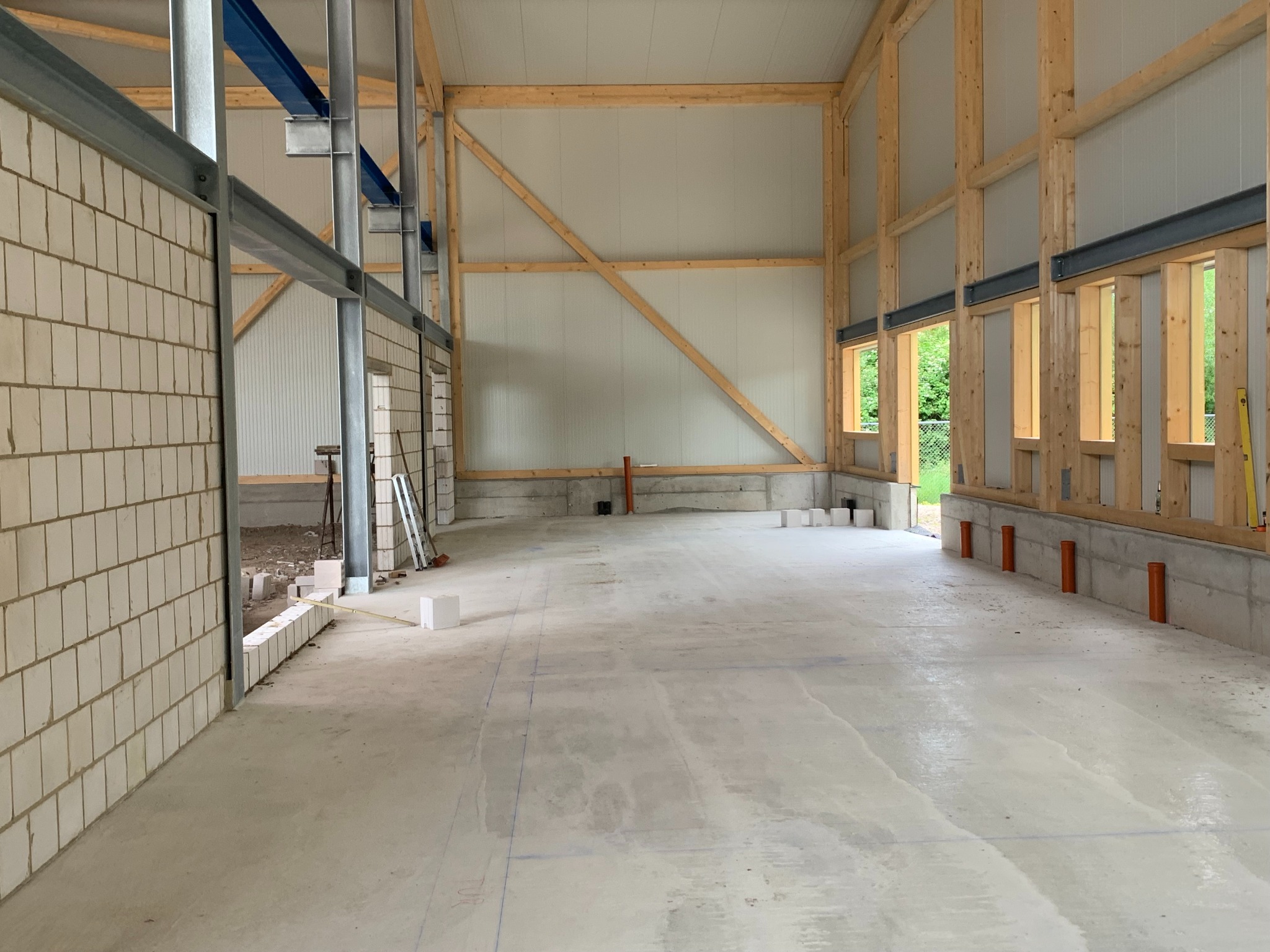 Wnętrze surowego domu murowanego w trakcie budowy, widoczne ściany z betonu komórkowego, drewniana konstrukcja dachu i stalowe belki, świeży beton na podłodze z zaznaczonymi liniami.