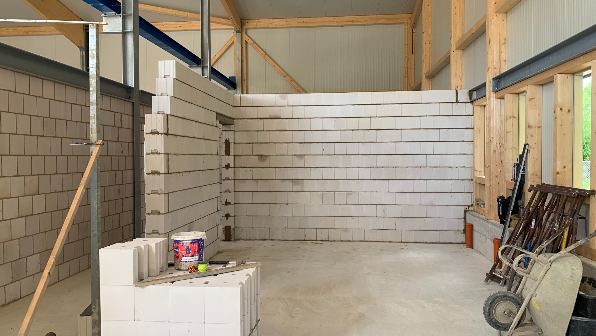 Wnętrze w trakcie budowy z białych bloczków, widoczne narzędzia budowlane, surowe drewniane elementy konstrukcyjne dachu i ścian, wózek budowlany z narzędziami w rogu.