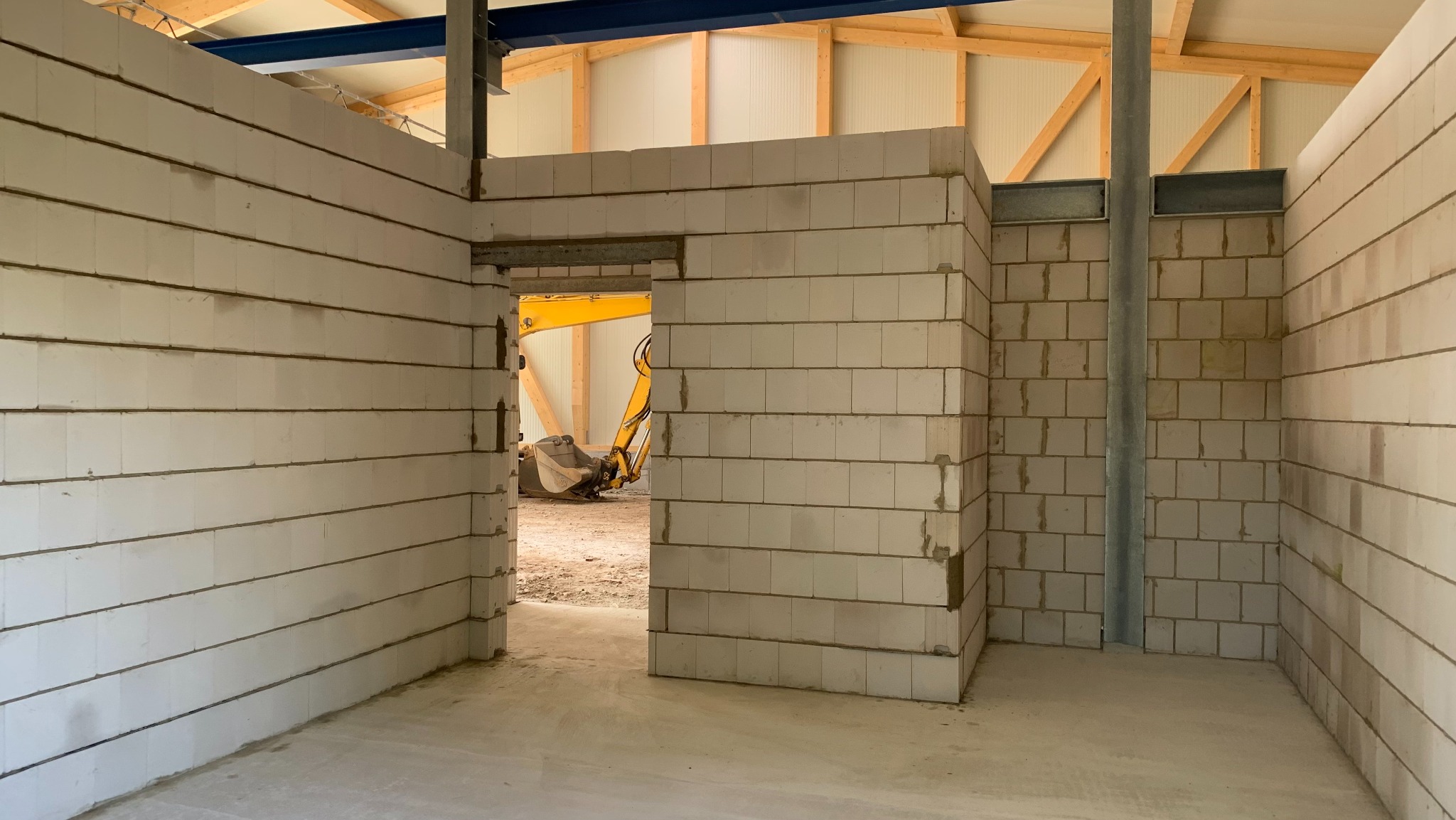 Wnętrze w trakcie budowy z białych bloczków, widoczny żółty sprzęt budowlany przez otwór drzwiowy, konstrukcja dachu z drewna i metalu.