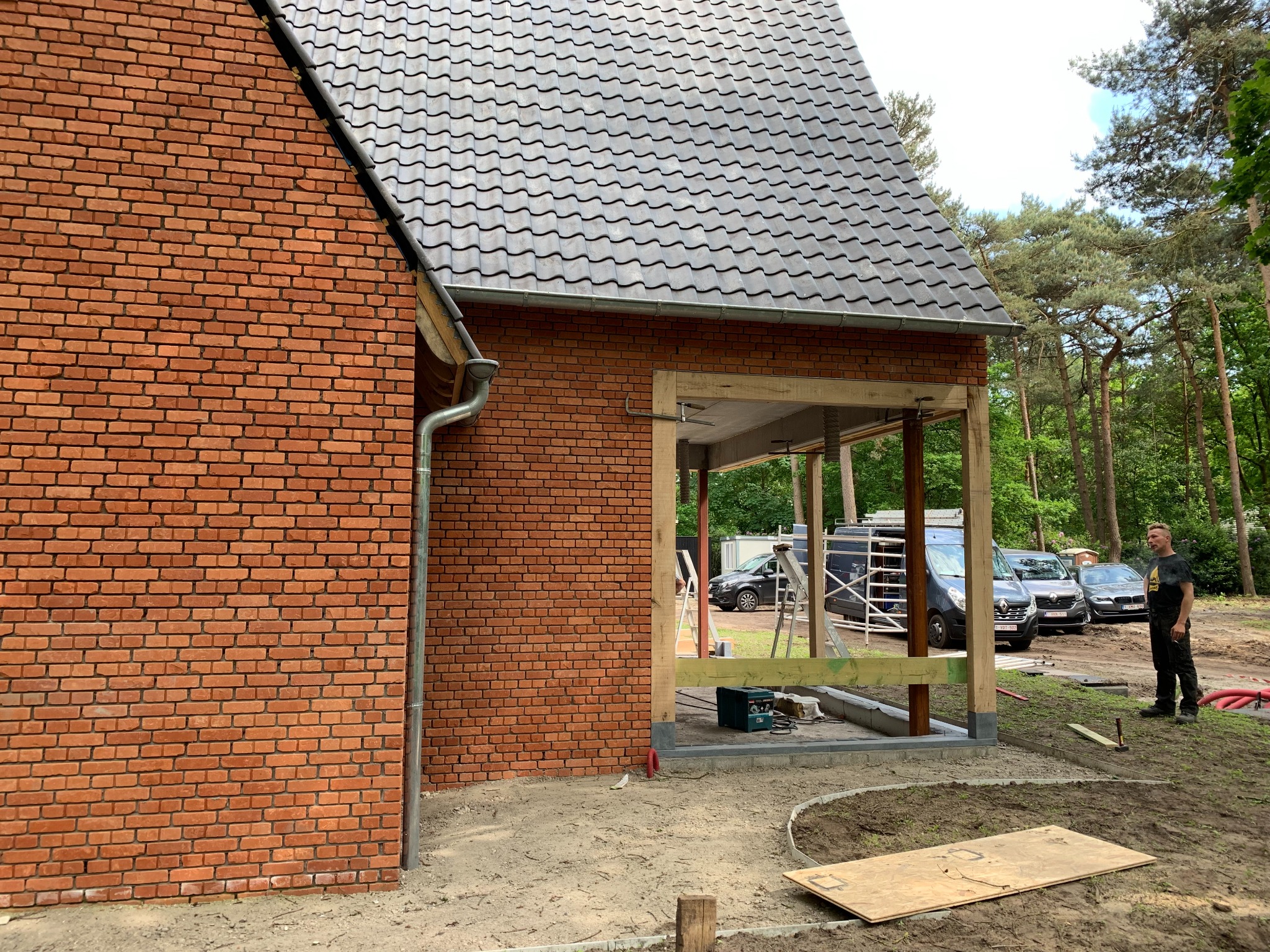 Częściowy widok murowanego domu z cegły z dobudowywaną drewnianą konstrukcją zadaszenia; widoczne narzędzia i samochody w tle.