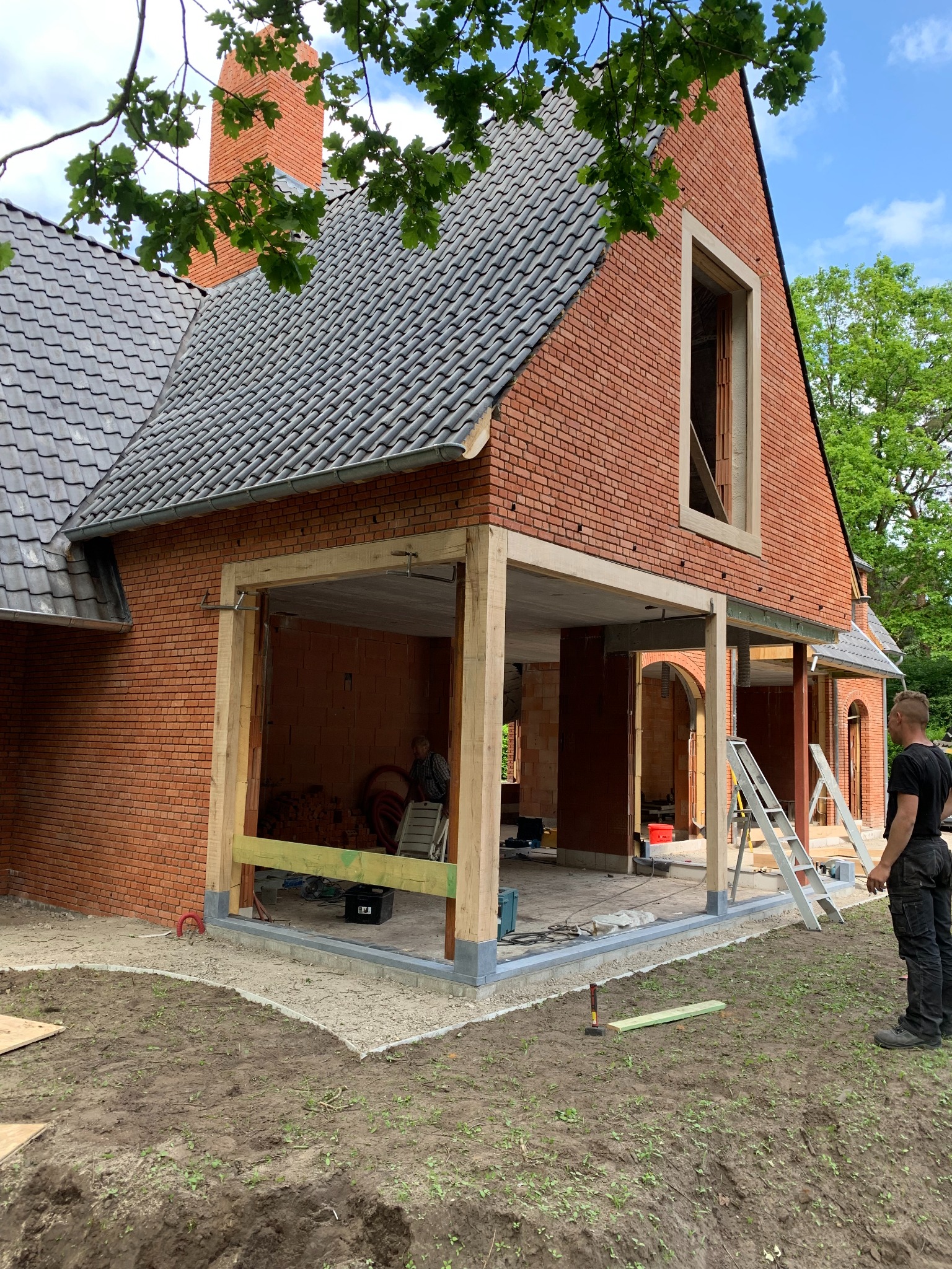 Budowa domu z czerwonej cegły z widocznym fragmentem dachu z szarej dachówki, drewnianą konstrukcją werandy i pracownikiem obserwującym postęp prac.
