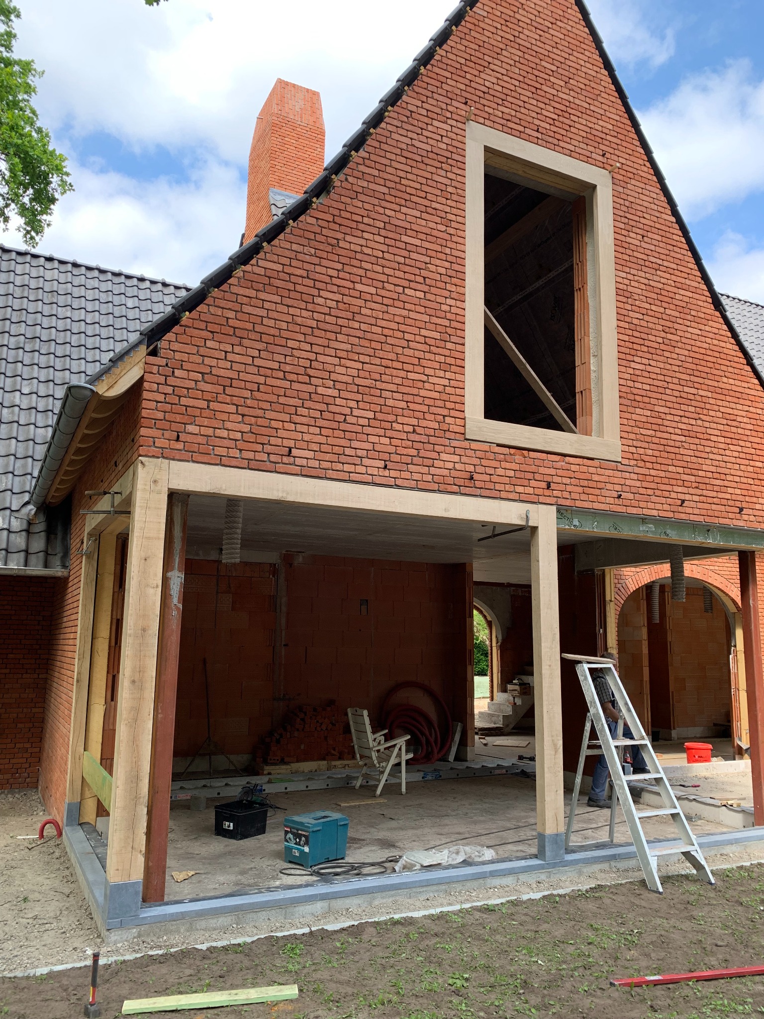 Budowa domu z czerwonej cegły w trakcie realizacji, widoczne drewniane podpory i rusztowanie, fragment dachu z szarej dachówki, drabina oparta o ścianę, narzędzia budowlane w tle, nieukończony komin.