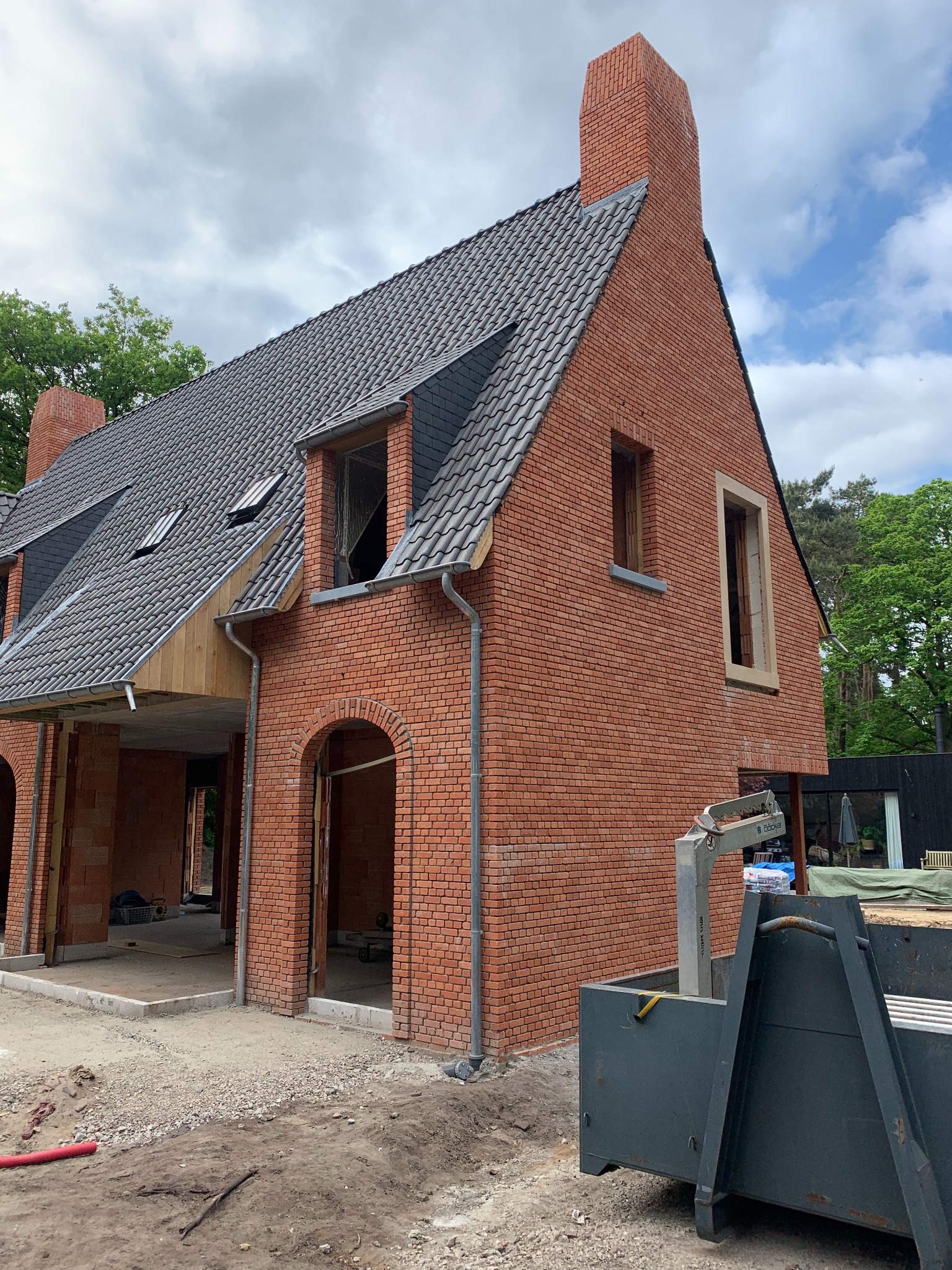 Surowy, dwukondygnacyjny dom z czerwonej cegły z ciemnoszarą dachówką, widoczne niezamontowane okna, obok kontener na gruz, w tle drzewa.