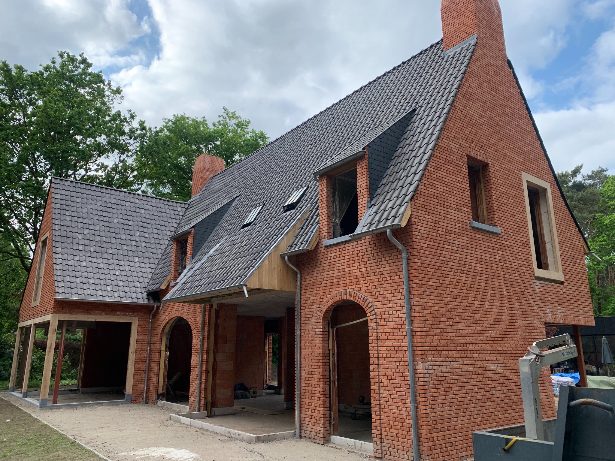 Surowy, ceglany dom w trakcie budowy z widocznymi elementami konstrukcyjnymi, szarym dachem i fragmentem lasu w tle.