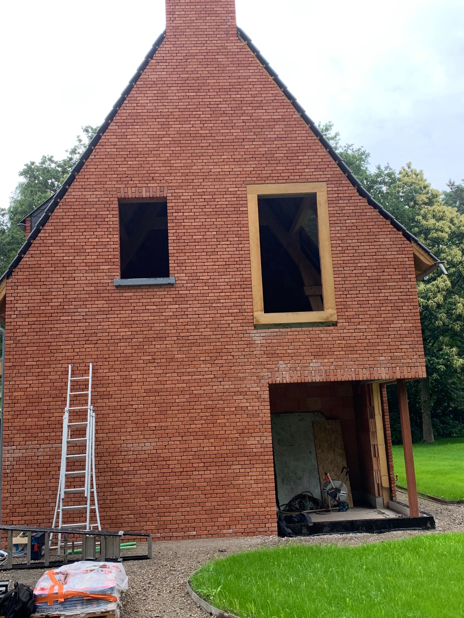 Wznoszony dom z czerwonej cegły z widoczną konstrukcją okienną i fragmentem dachu, na tle zieleni, z drabiną opartą o ścianę i materiałami budowlanymi na palecie.