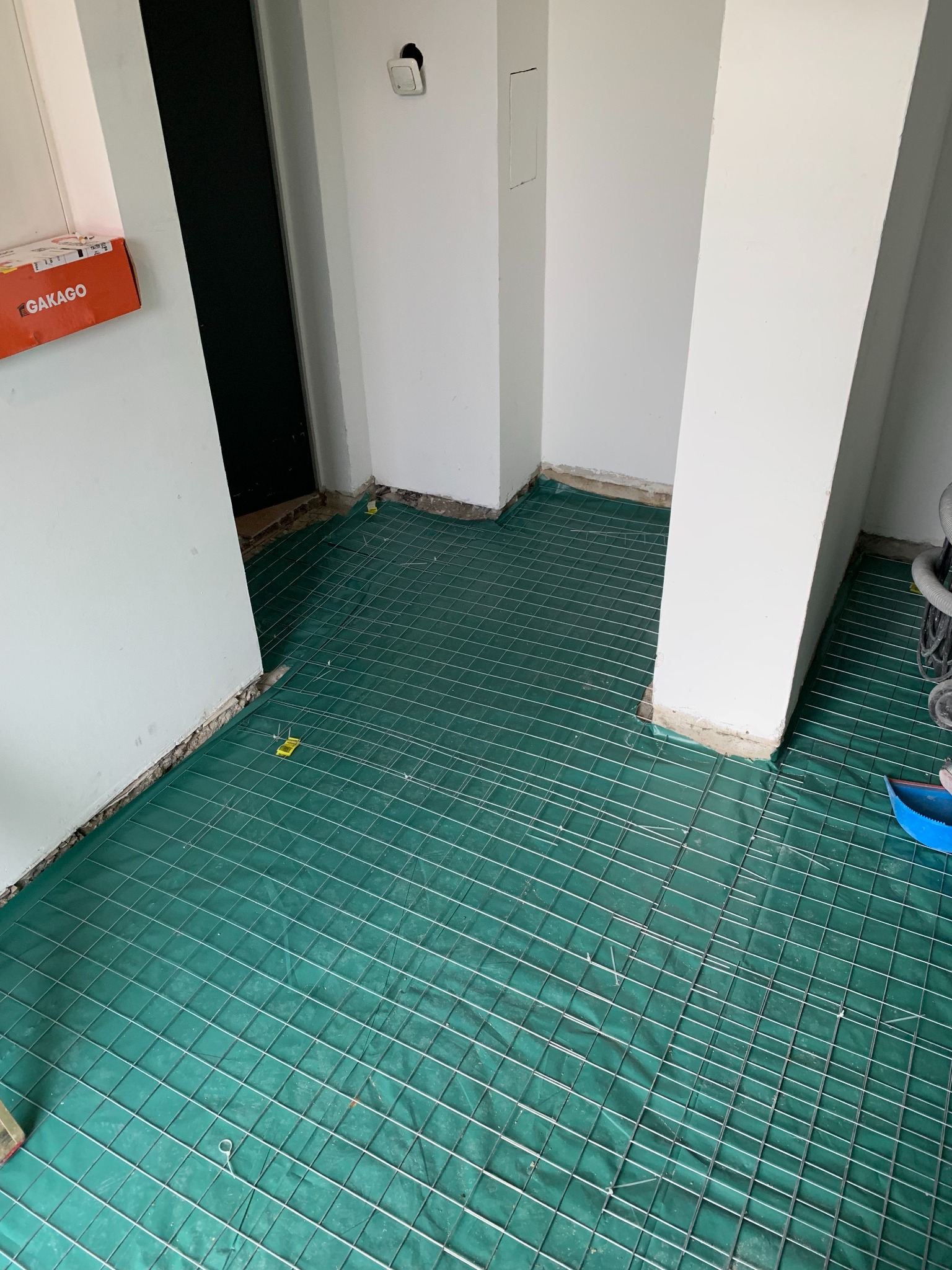 Przygotowanie podłoża pod ogrzewanie podłogowe: siatka z drutu na zielonej folii, widoczne elementy mocujące i fragment ściany z puszką elektryczną.