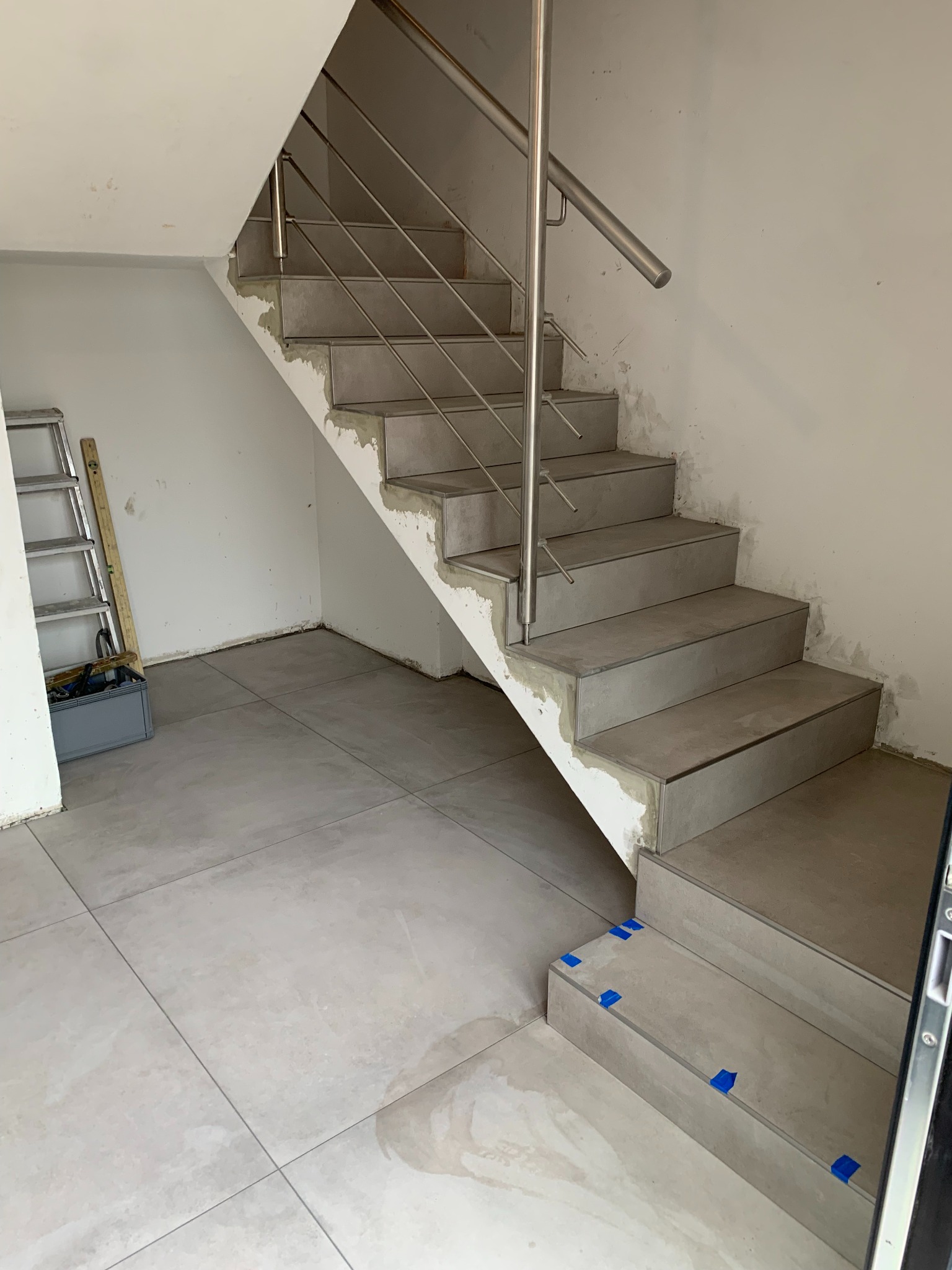 Montaż płytek na schodach z metalową balustradą, widoczne ślady zaprawy i niebieska taśma zabezpieczająca, drabina w tle.
