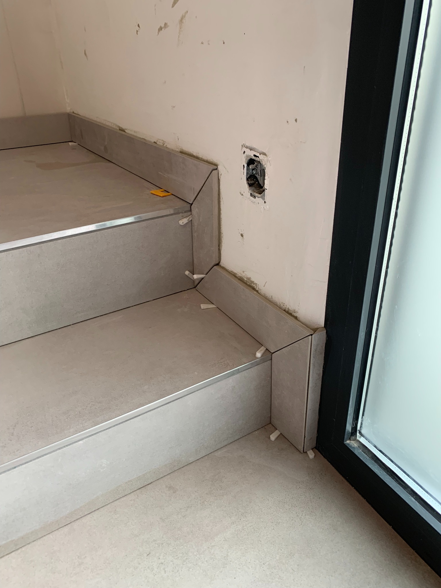 Wykończenie schodów płytkami ceramicznymi w szarym odcieniu, z plastikowymi klinami dystansowymi, widoczny fragment ściany z puszką elektryczną i czarna rama okienna.