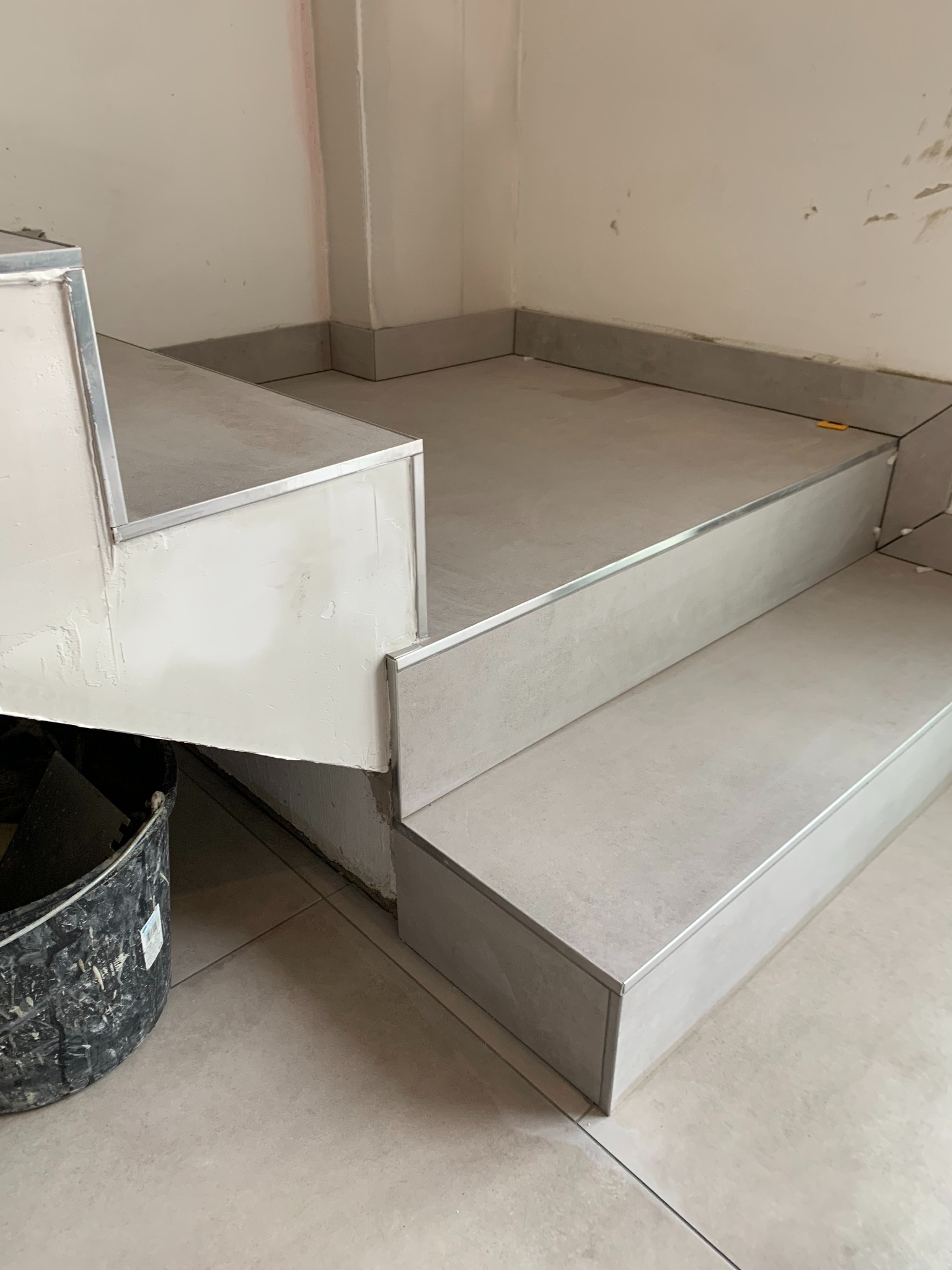 Wykończenie schodów płytkami ceramicznymi z metalowymi listwami, widoczny fragment ściany z pozostałościami zaprawy i wiadro z narzędziami.