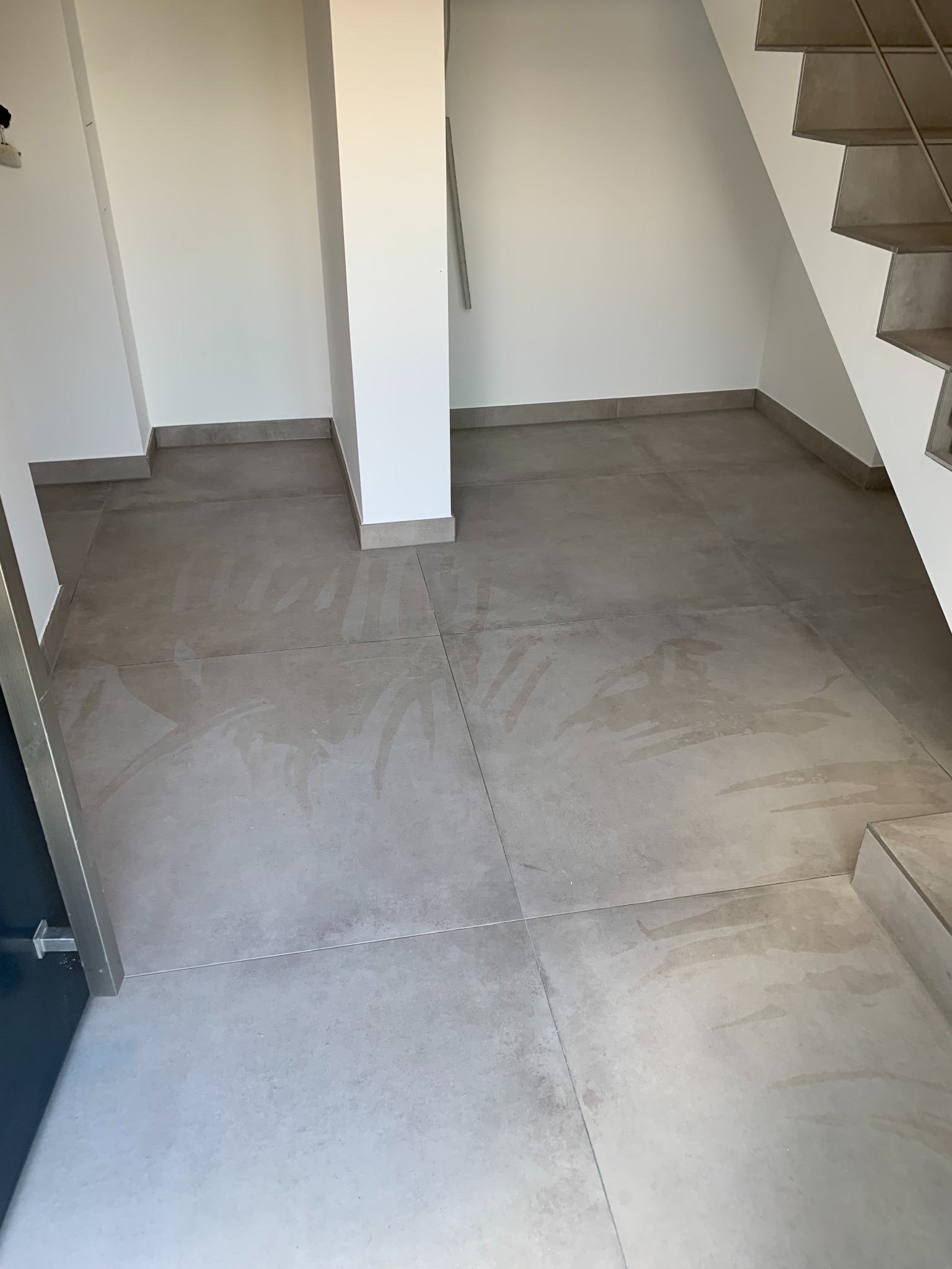 Szare płytki podłogowe w nowym domu, widoczne ślady po myciu, białe ściany i fragment schodów z szarą balustradą.