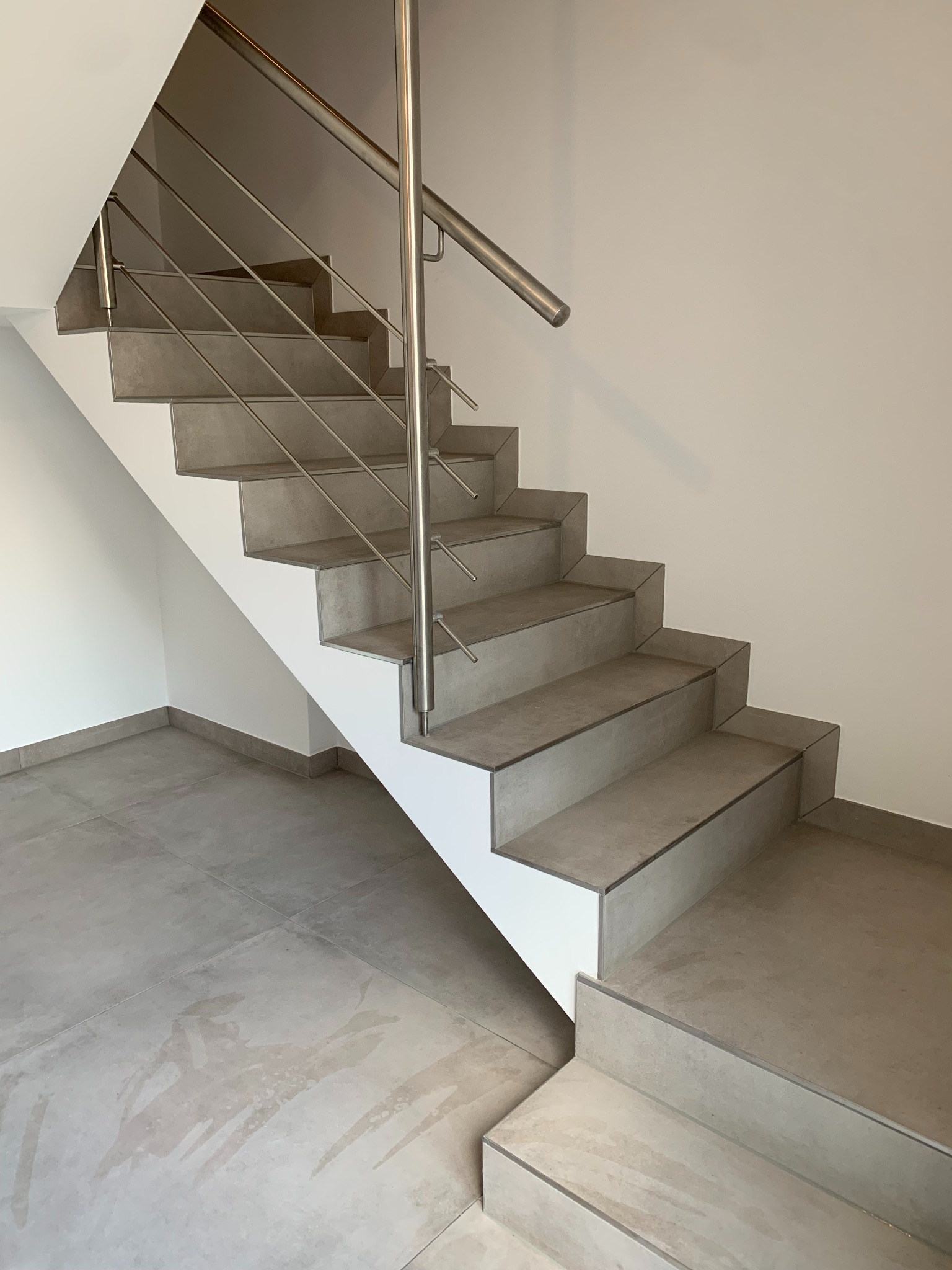 Nowoczesne schody wewnętrzne z szarych płytek ceramicznych z metalową balustradą, widoczne z dołu, minimalistyczne wykończenie ścian i podłogi w odcieniach szarości.