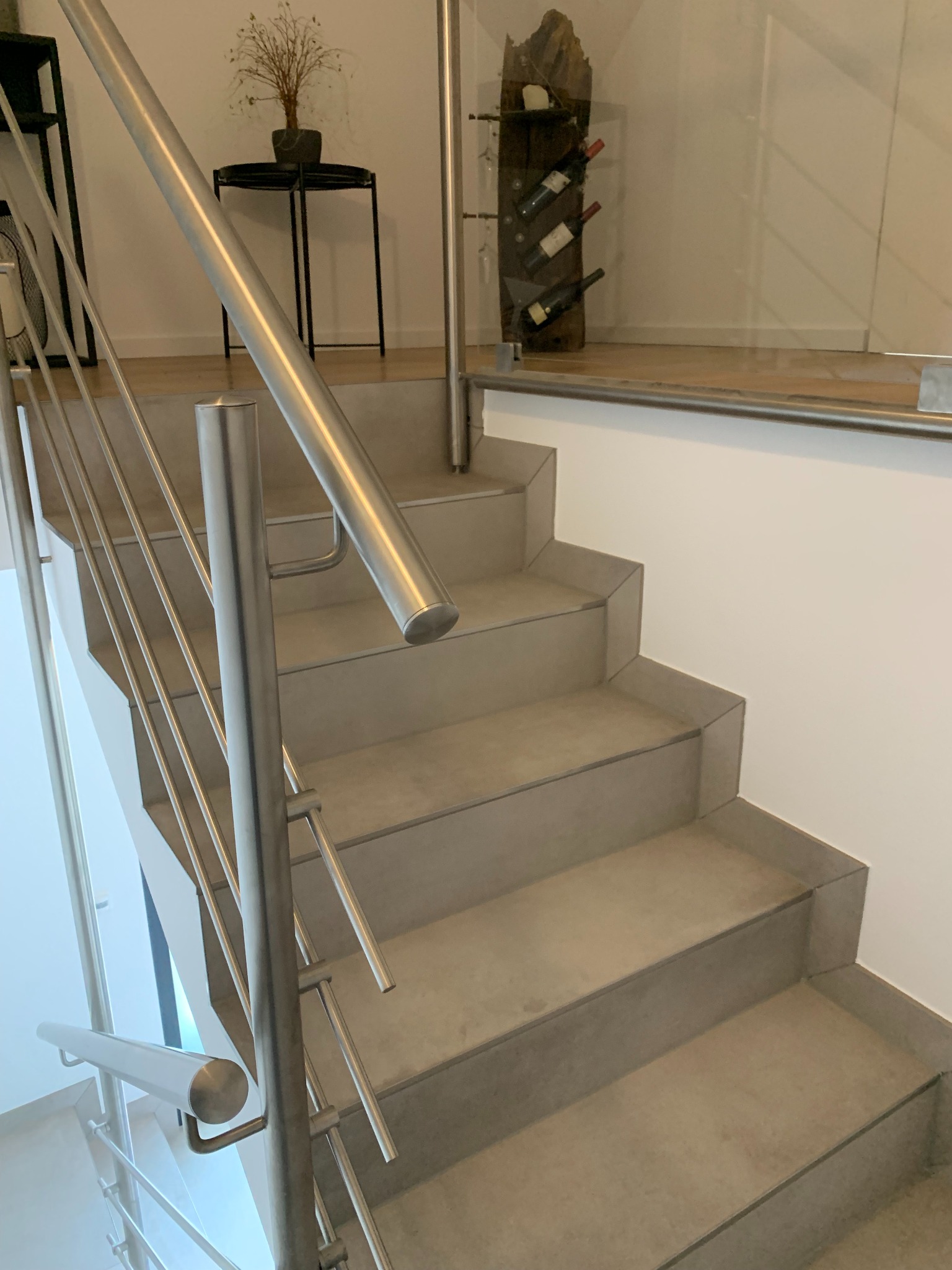 Schody wewnętrzne wykończone płytkami w odcieniach szarości, z nowoczesną balustradą ze stali nierdzewnej i szklanym panelem, widoczny element dekoracyjny - stojak na wino z butelkami.