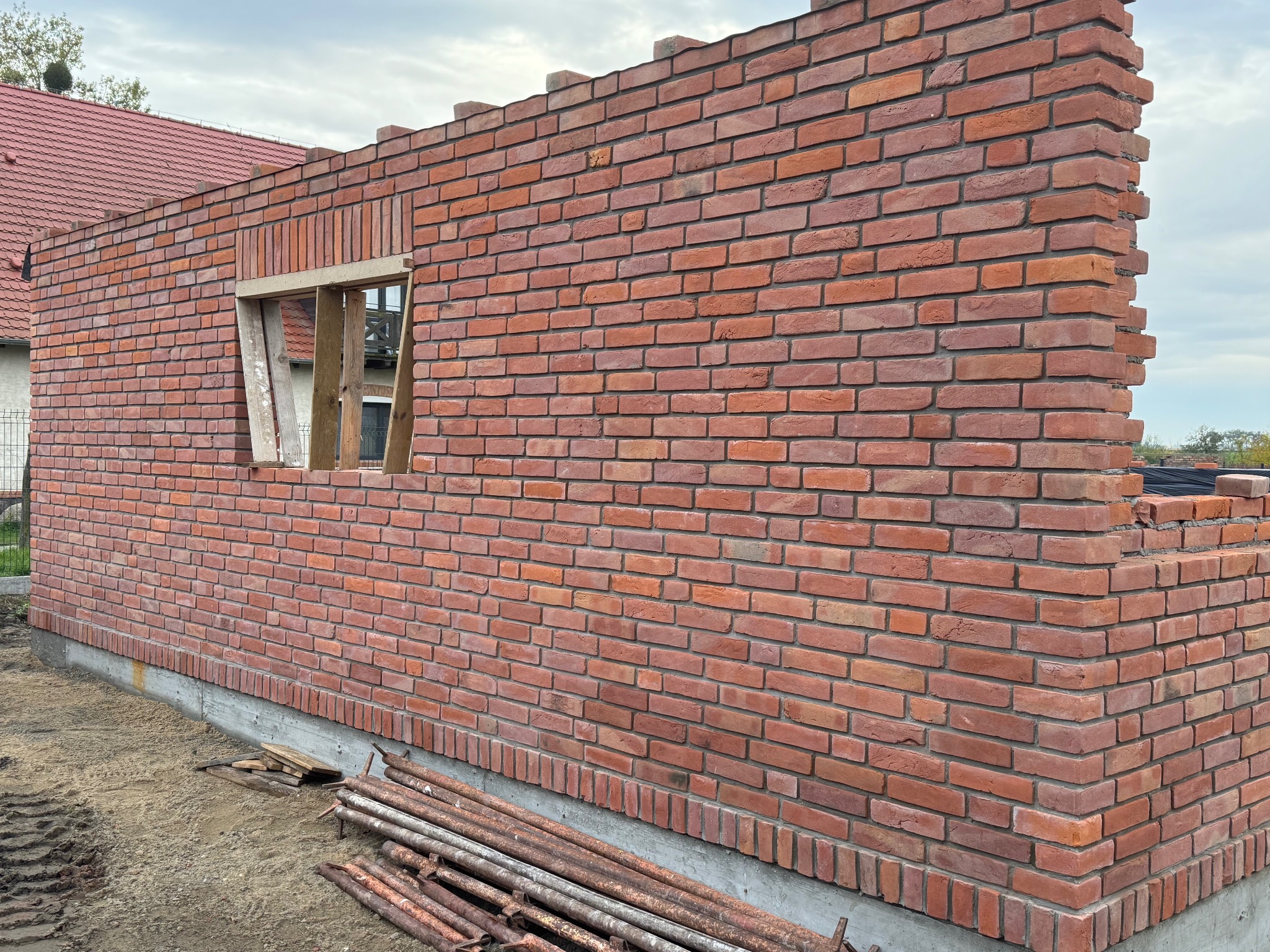 Wznoszenie ściany z czerwonej cegły z otworem okiennym, widoczne rusztowania i fragment dachu budynku w tle. Prace murarskie w toku.