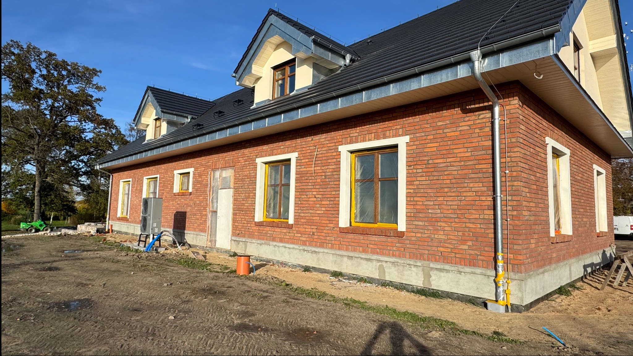 Ceglany dom z czarnym dachem i żółtymi ramami okiennymi w trakcie budowy, widoczne instalacje i teren wokół domu. Częściowo wykończona elewacja.
