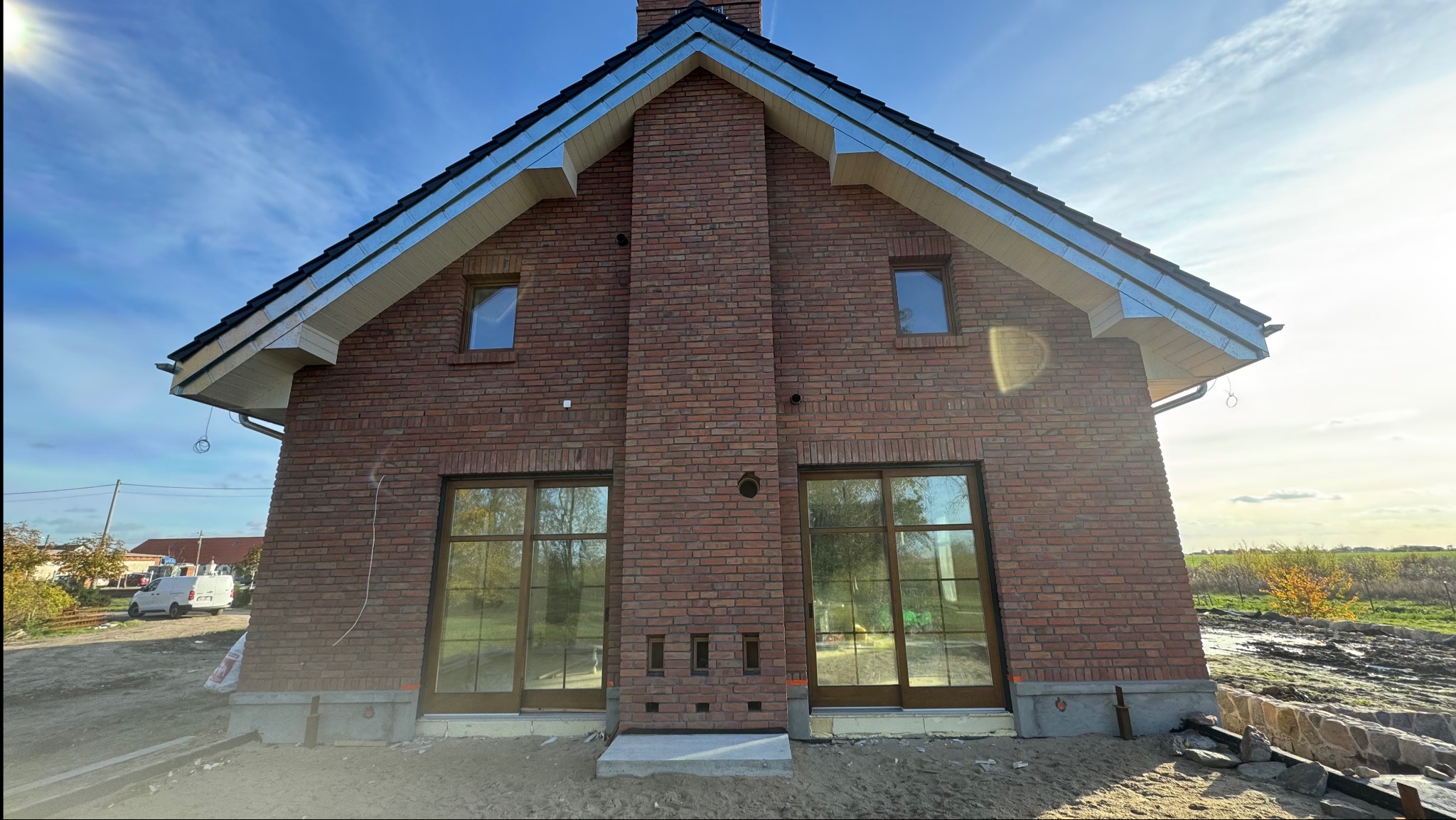 Nowoczesny dom murowany z czerwonej cegły, z dużymi oknami i kominem. Widoczne detale wykończenia dachu i elewacji. W tle błękitne niebo i częściowo zagospodarowany teren.