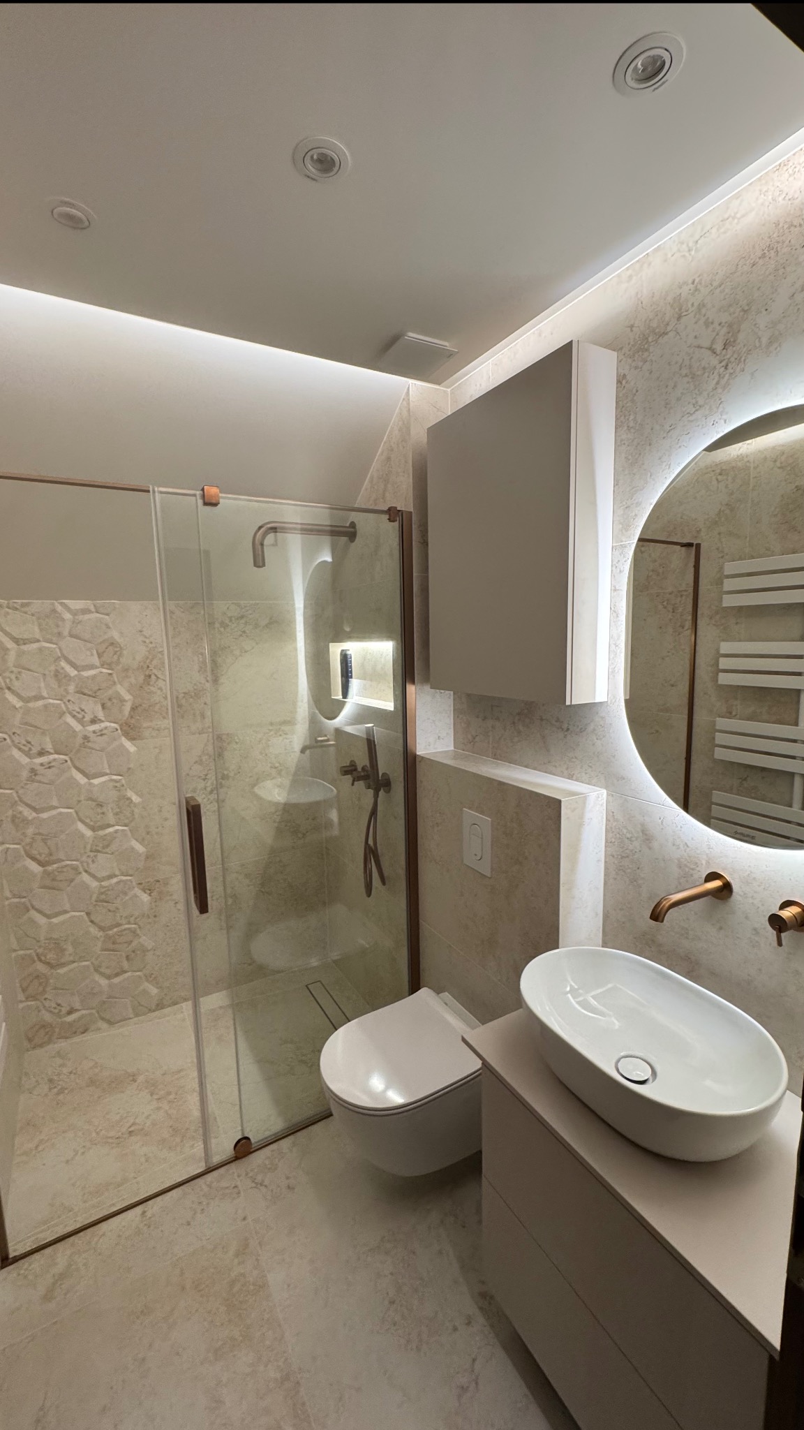 Nowoczesna łazienka z kabiną prysznicową typu walk-in, umywalką nablatową i okrągłym lustrem z podświetleniem LED. Beżowe płytki imitujące kamień na ścianach i podłodze.