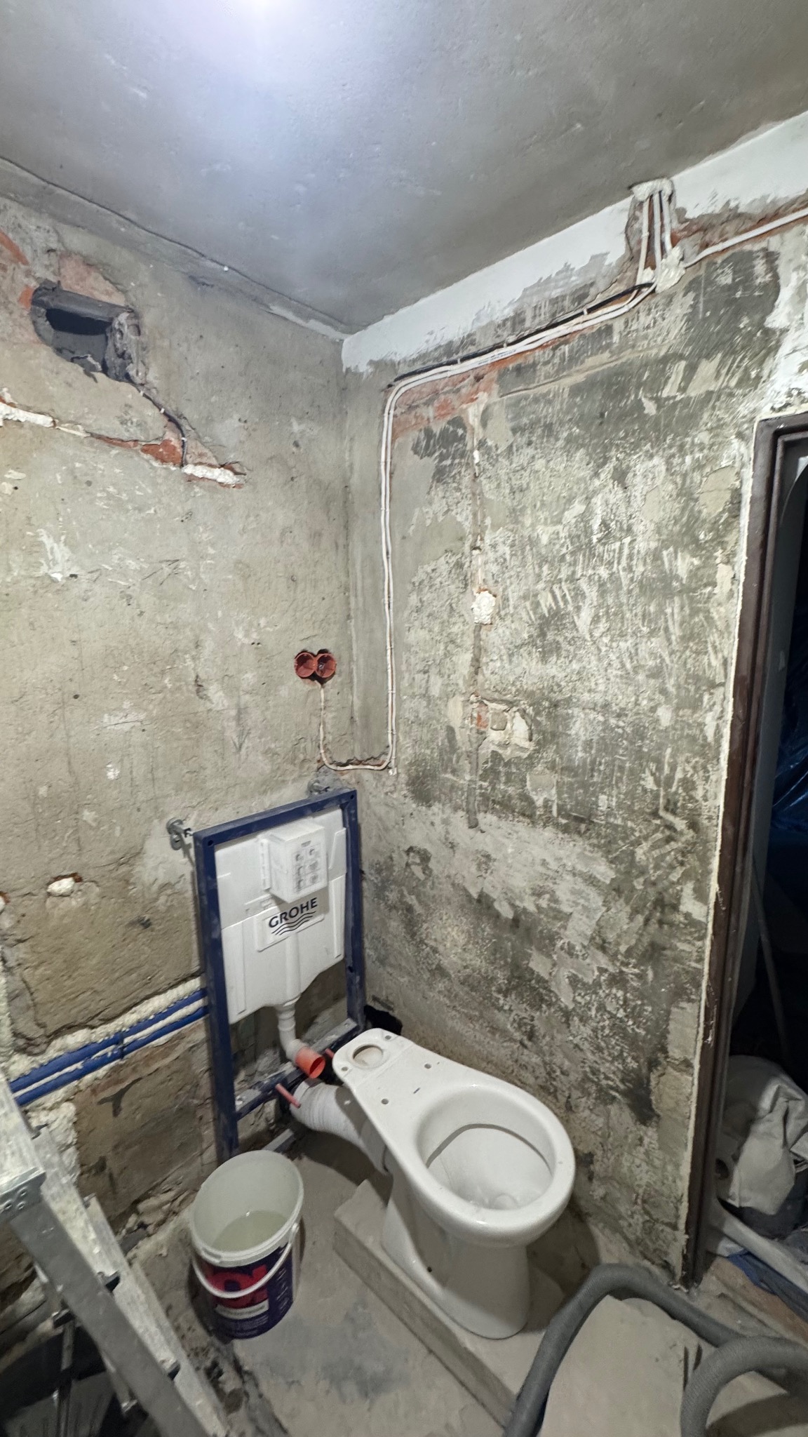 Surowa łazienka w trakcie remontu: widoczne instalacje, stelaż podtynkowy Grohe, częściowo zamontowana toaleta, ściany w stanie deweloperskim z odsłoniętą instalacją elektryczną.