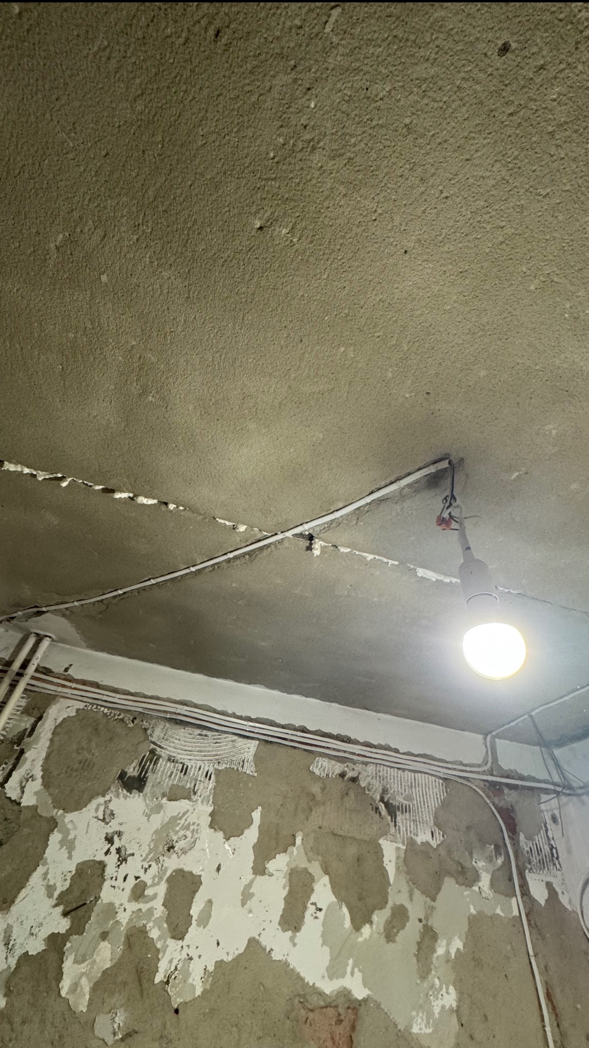 Ściana w trakcie skuwania tynku, widoczne instalacje elektryczne i rury. Sufit z widocznymi kablami i prowizorycznym oświetleniem. Etap przygotowania do remontu.