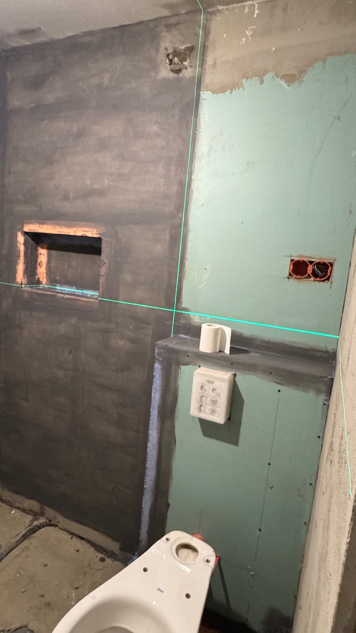 Niedokończona łazienka z surowymi ścianami, zielonymi płytami gipsowymi, laserem krzyżowym, wnęką i częściowo widoczną muszlą klozetową. Etap prac wykończeniowych.