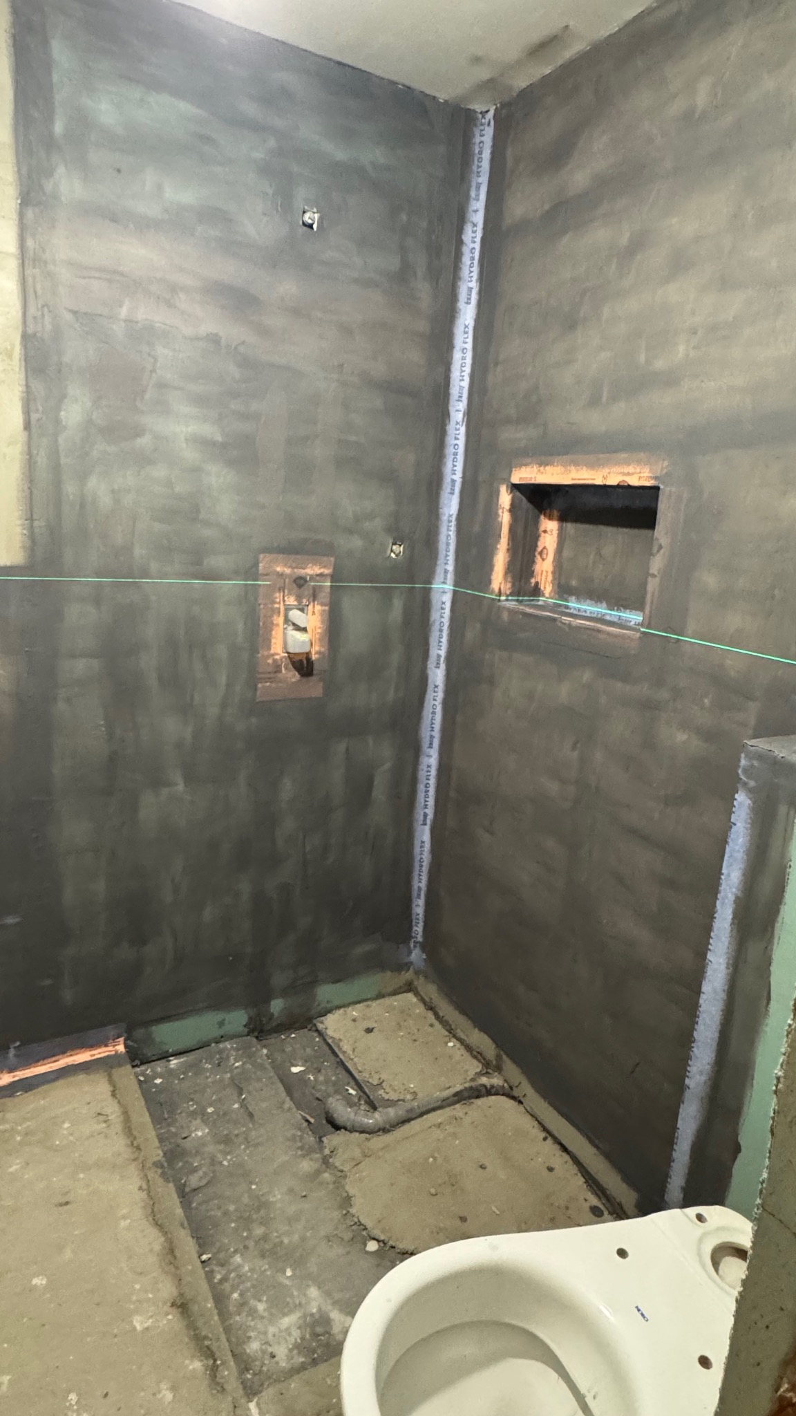 Przygotowana do wykończenia łazienka z szarymi ścianami, widoczną instalacją i częściowo zamontowaną toaletą. W tle otwór w ścianie.