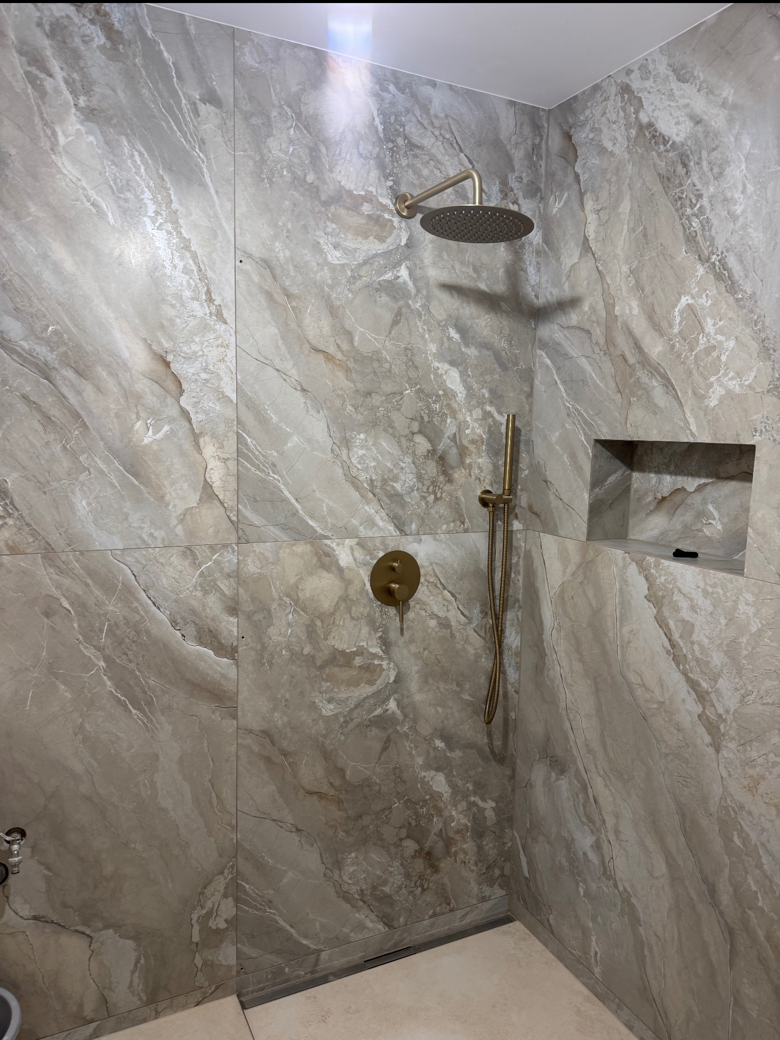 Wykończona kabina prysznicowa z beżowymi płytkami imitującymi marmur, złotą armaturą i wnęką na kosmetyki. Minimalistyczny design, eleganckie wykończenie.