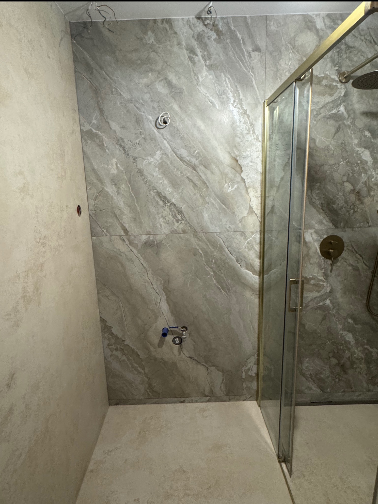 Wykończona łazienka z kabiną prysznicową typu walk-in, wyłożona płytkami imitującymi marmur w odcieniach szarości i beżu. Widoczne elementy instalacji wodnej i elektrycznej.