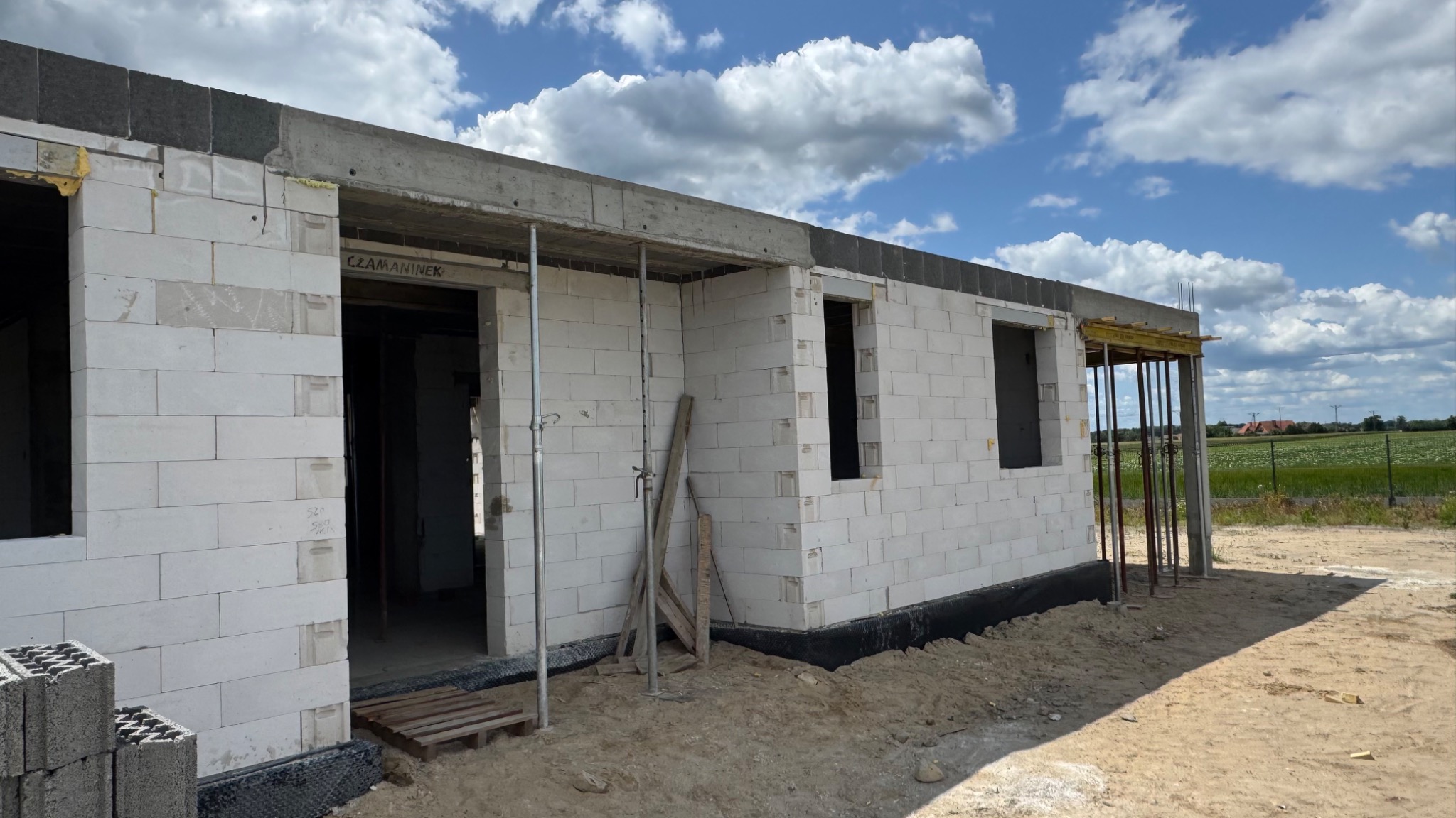 Wznoszenie ścian parterowego domu z białych bloczków, widoczne otwory okienne i drzwiowe, szalunki stropowe. W tle błękitne niebo z chmurami i zielone pola.