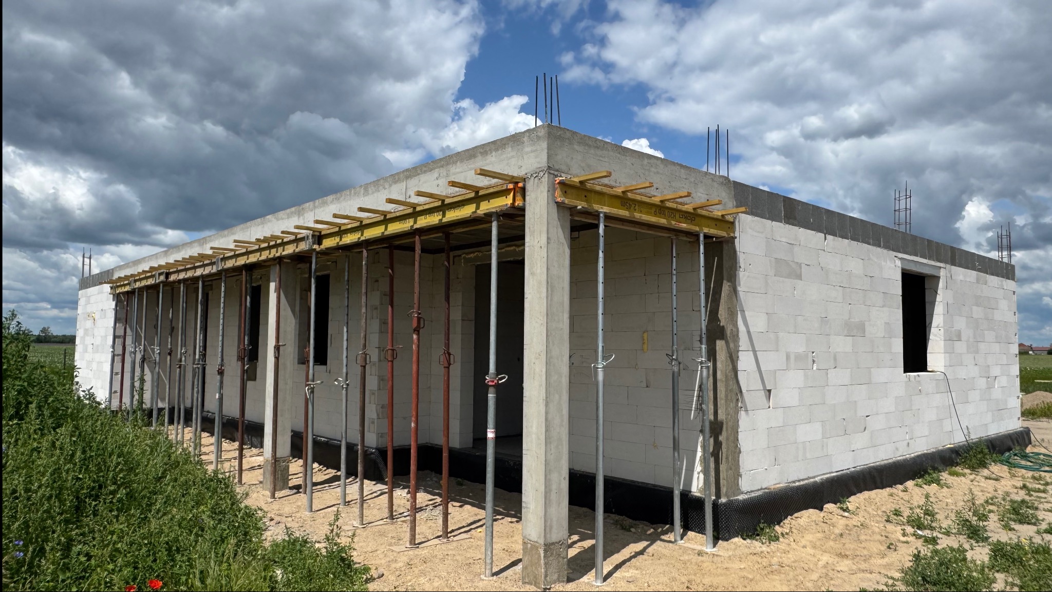 Surowy stan domu z betonu komórkowego, z widocznymi podporami stropu i zbrojeniami, na tle błękitnego nieba z chmurami i zielenią w tle.