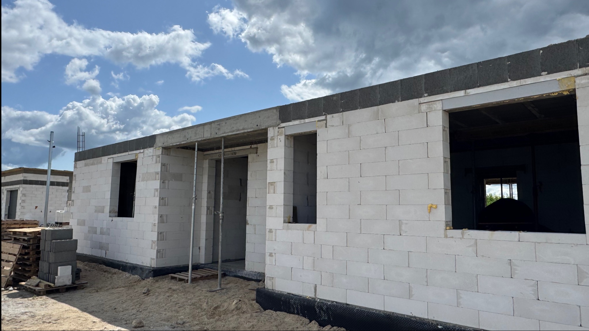 Budowa domu z białych bloczków w Zagórowie. Widoczne ściany w stanie surowym otwartym, okna bez szyb, palety z materiałami budowlanymi. Niebo z chmurami w tle.