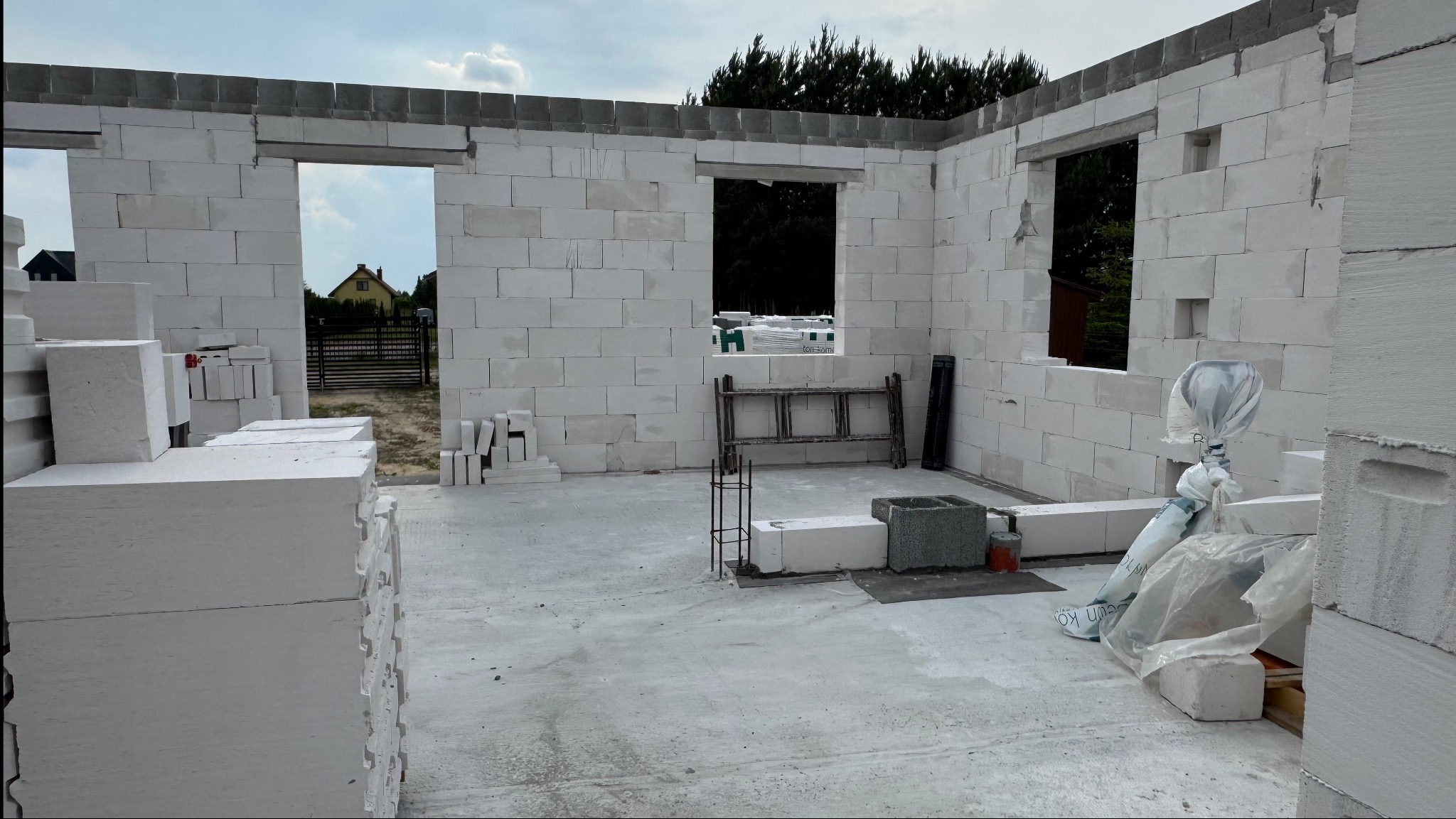 Wnętrze surowego domu z białych bloczków, widoczne otwory okienne i drzwiowe, przygotowane do dalszych prac budowlanych. Na podłodze leżą materiały budowlane.